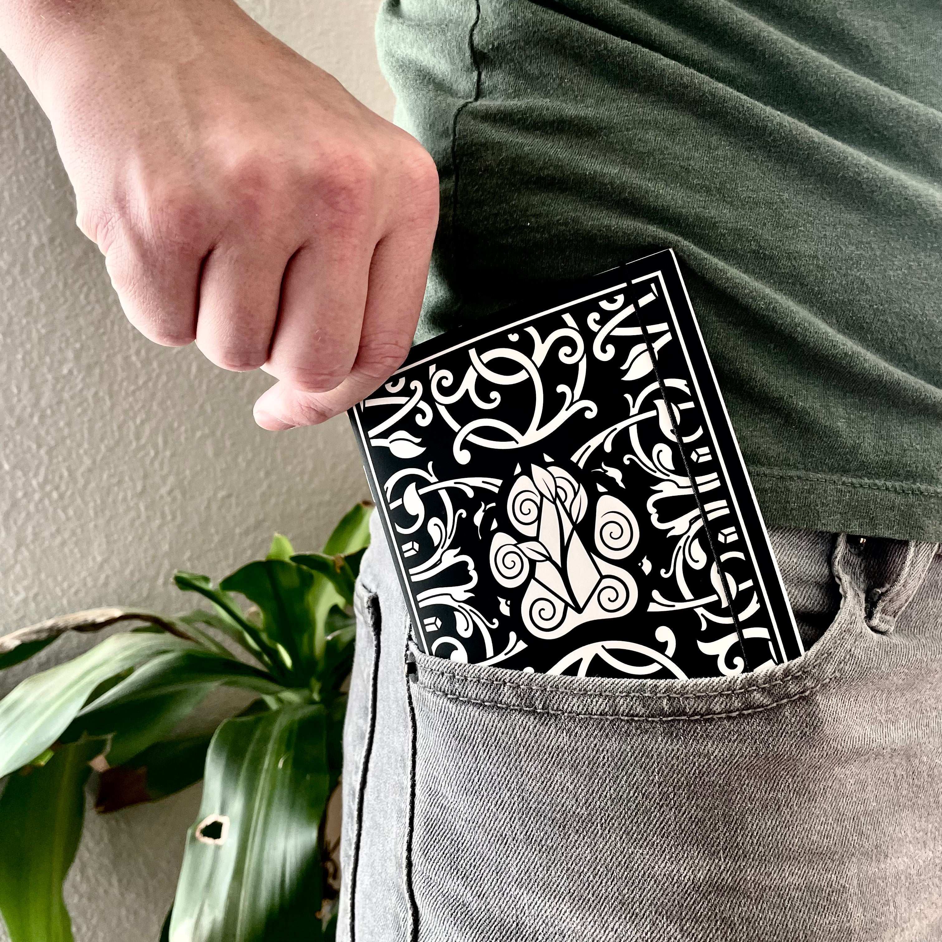 Class Cover 5E Character Journals - Black journal with intricate white fantasy designs on the cover, held in a person's gray jeans pocket.