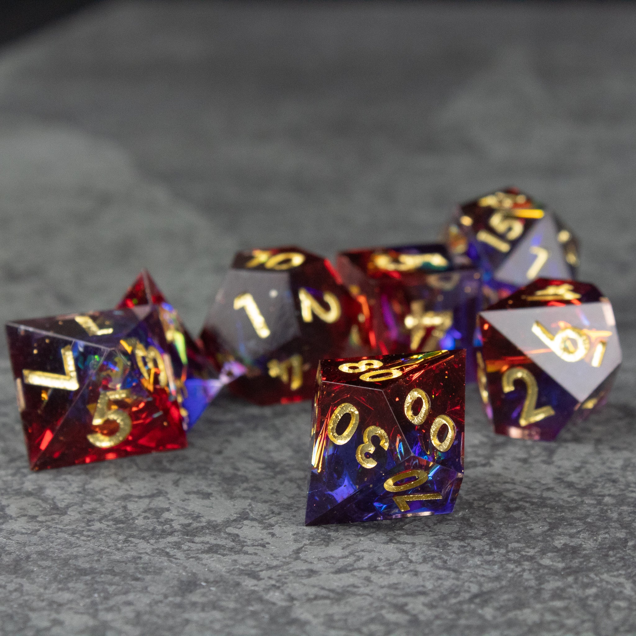 Electromancer Dice Set - Set of transparent blue and red sharp-edged polyhedral dice with gold numbers on gray surface.