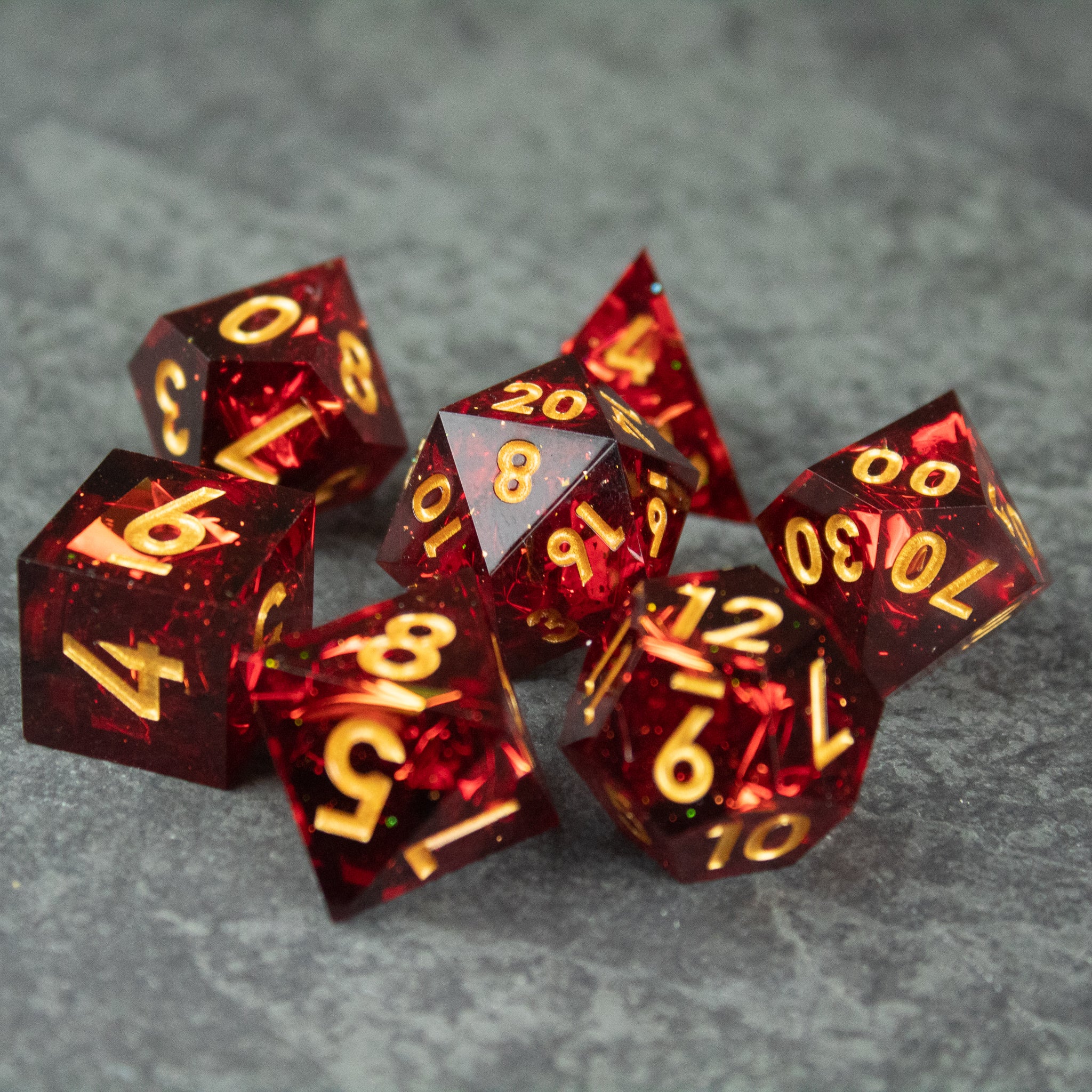 Ember Dice Set - Set of seven translucent red polyhedral dice with sharp edges and gold numbers on a gray textured surface.