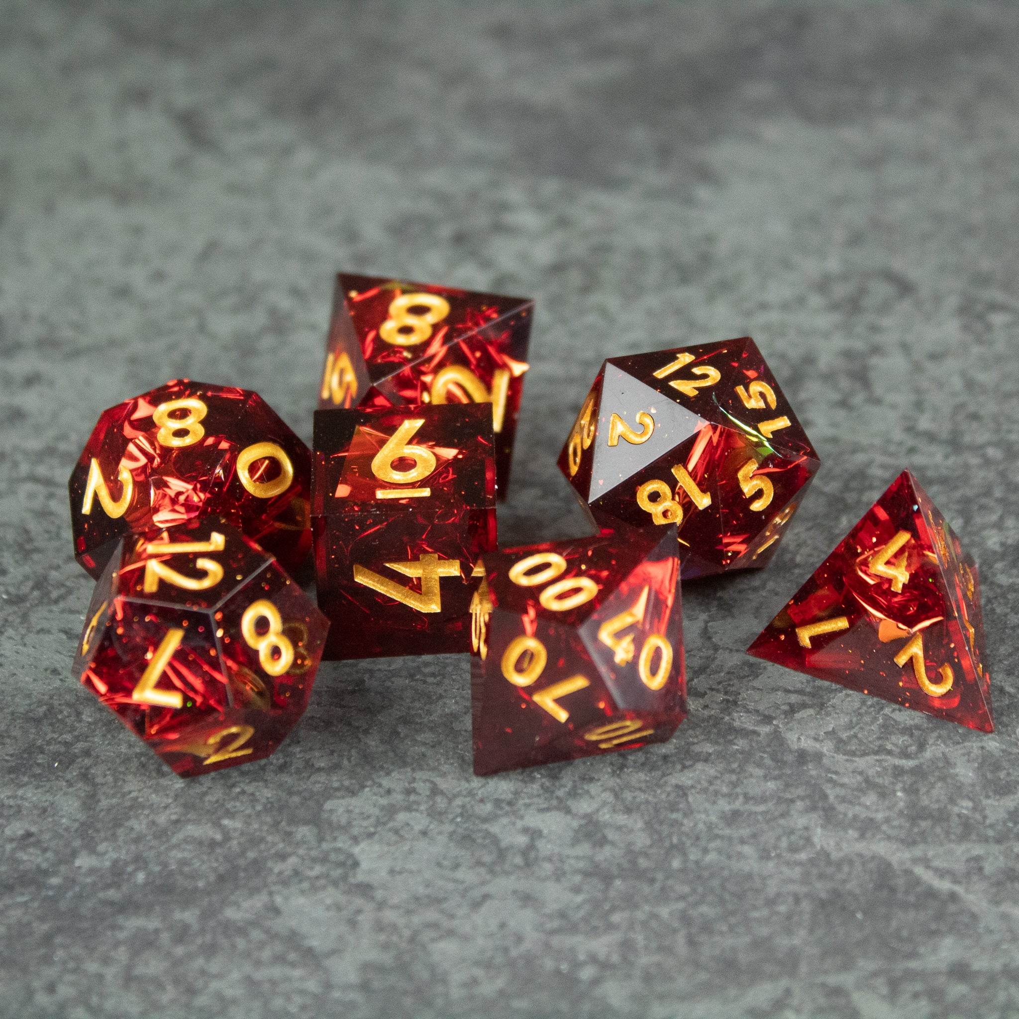 Ember Dice Set - Set of seven sharp-edged translucent red polyhedral dice with gold numbers on gray surface.