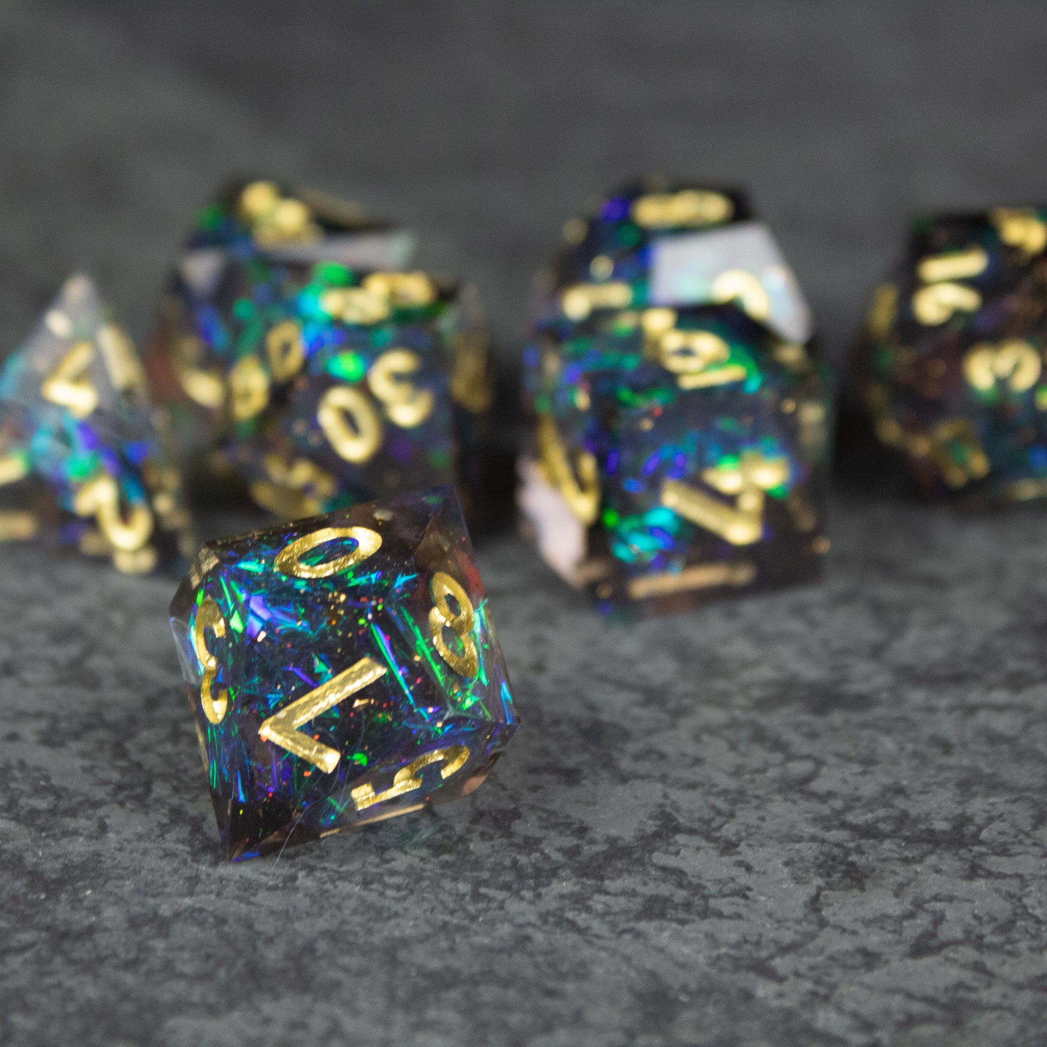 Galaxy Dice Set - Black sharp-edged polyhedral dice with shimmering blue and green galaxy-like patterns and gold numbering on a textured dark.