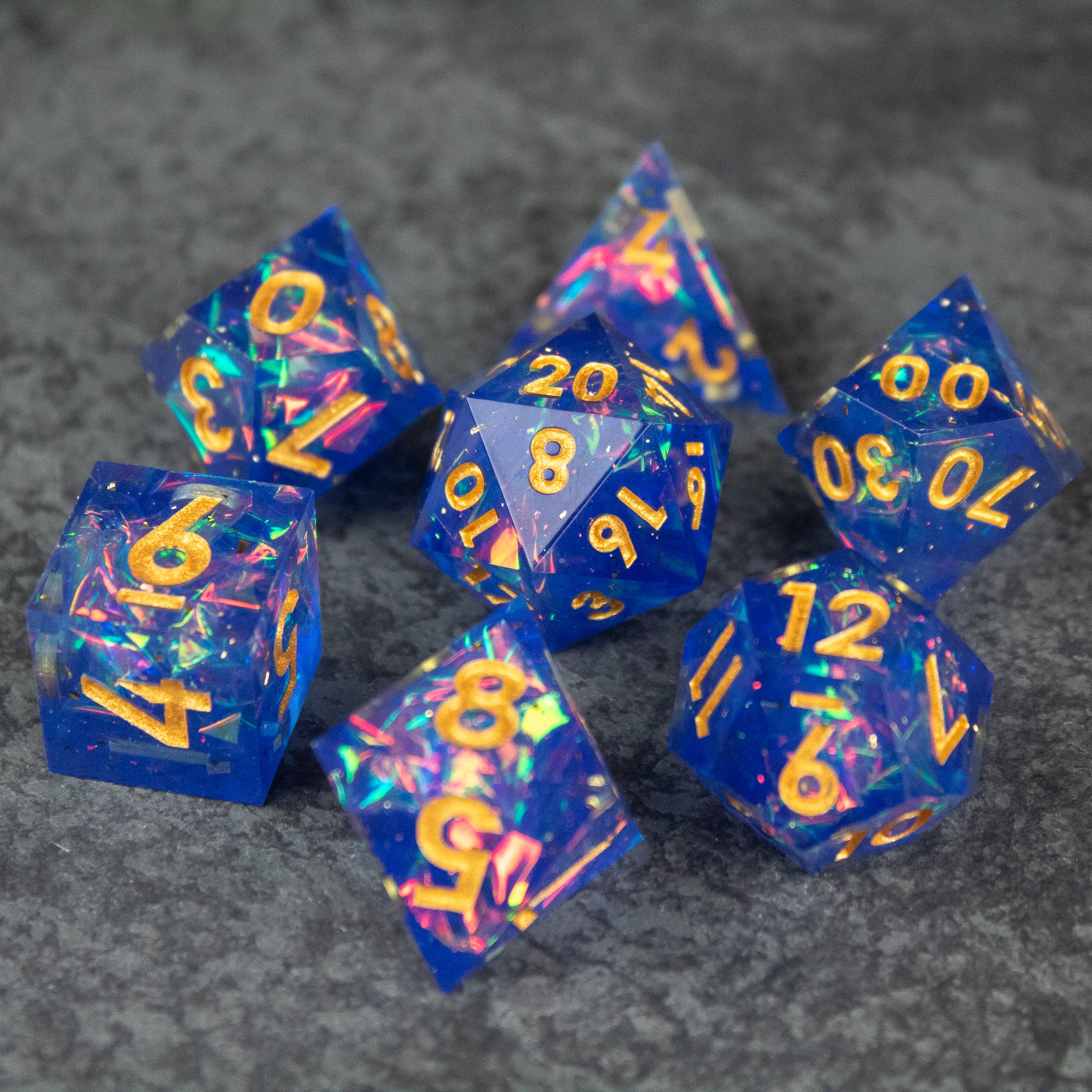 Arcana Dice Set - Set of seven blue sharp-edged dice with multicolored glitter and gold numbers on gray stone surface.