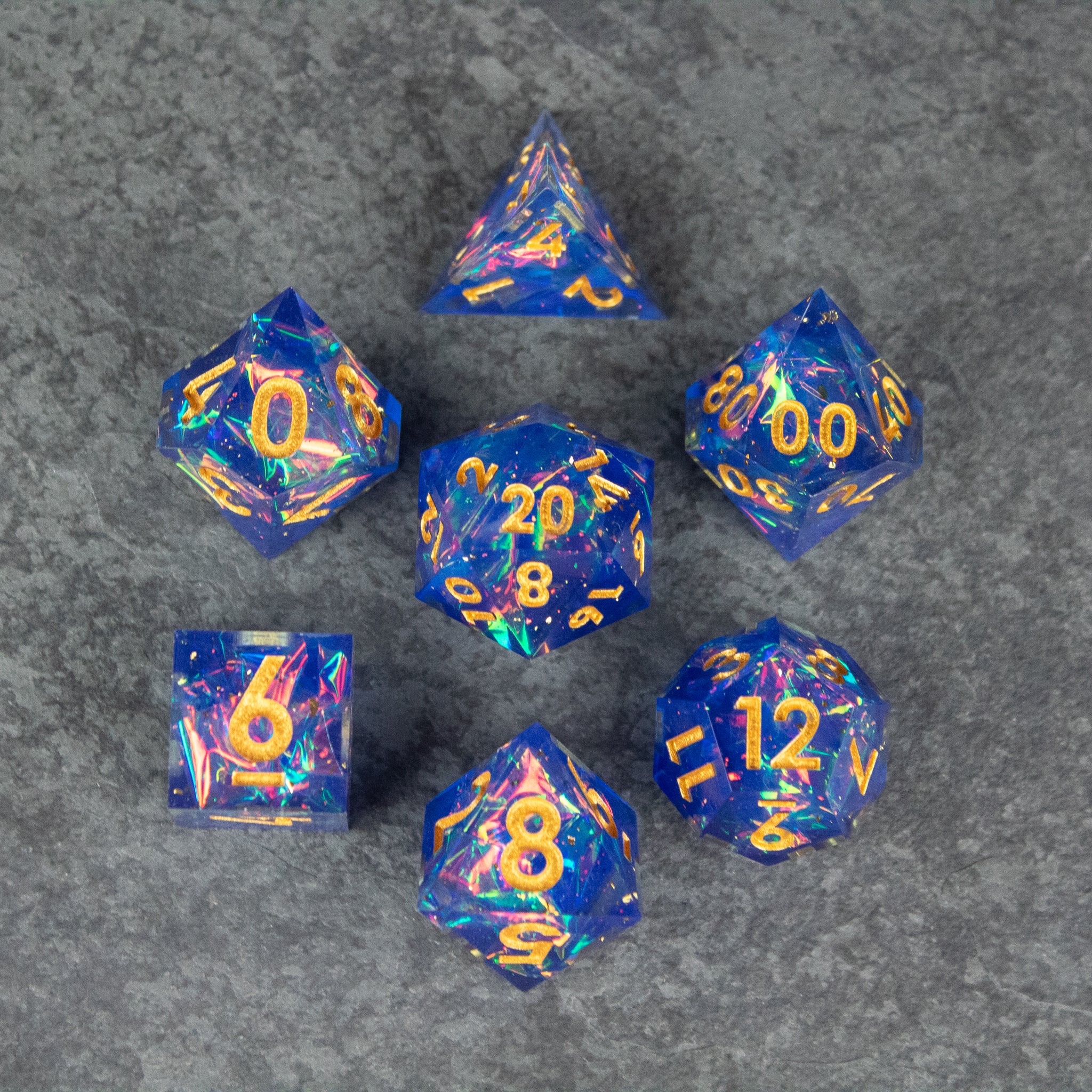 Arcana Dice Set - Blue sharp-edged polyhedral dice set with yellow numbers and gold swirling patterns on gray surface.