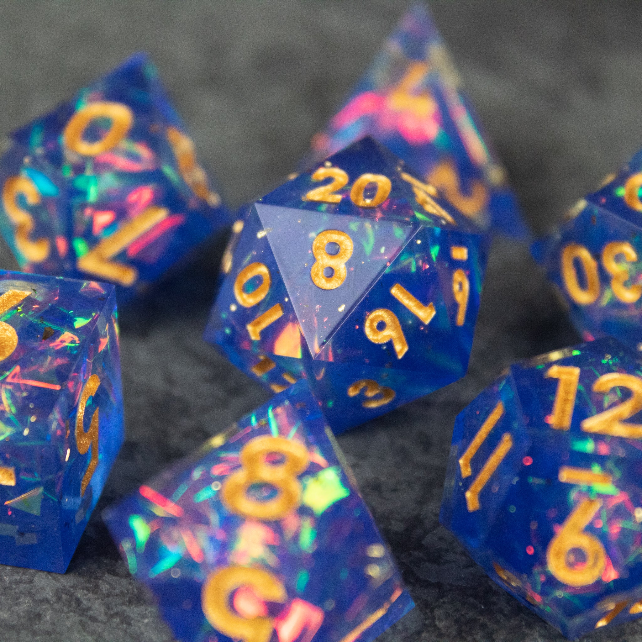 Arcana Dice Set - Set of translucent blue sharp-edged polyhedral dice with gold numbering and colorful iridescent flakes inside.
