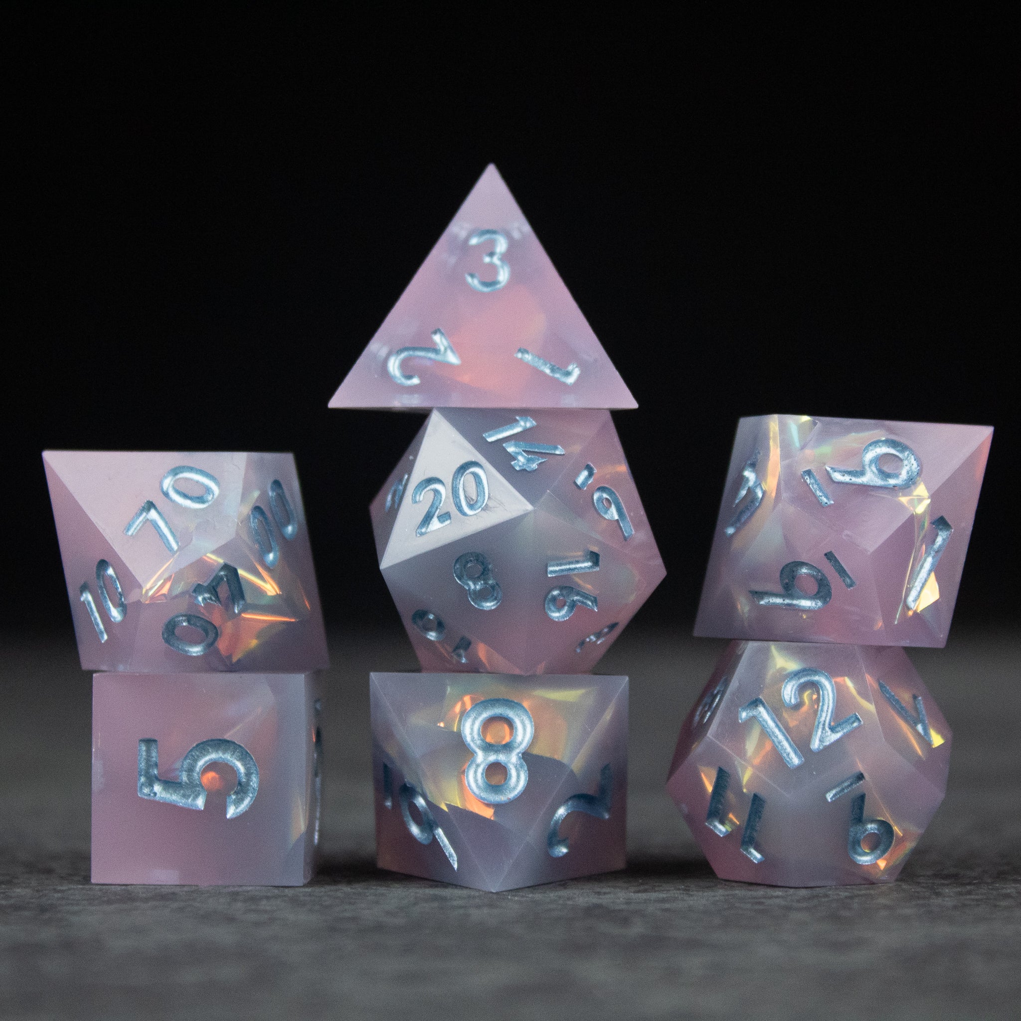 Moonshard Dice Set - Set of translucent pink sharp-edged dice with blue metallic numbers on gray surface.