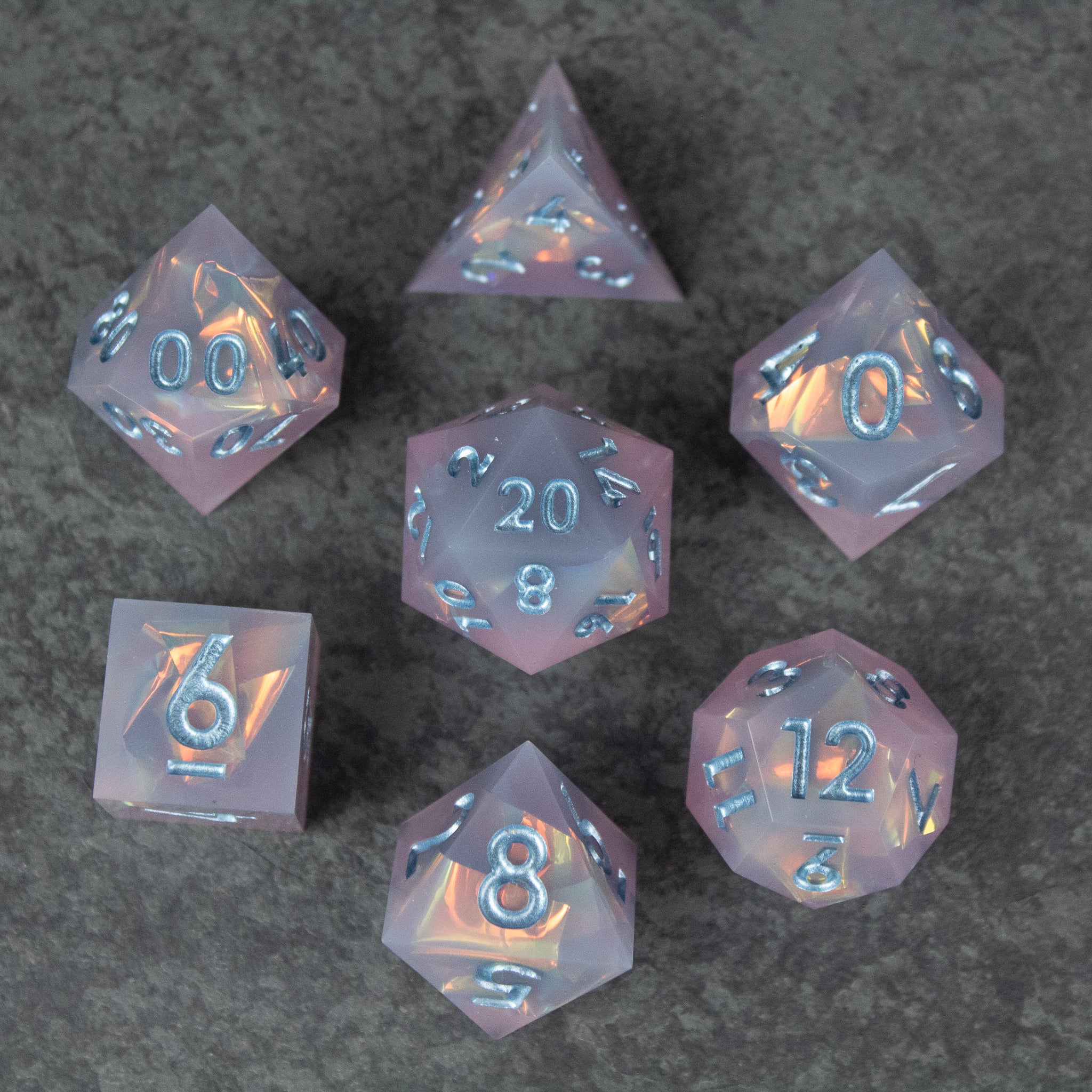 Moonshard Dice Set - Translucent pink seven-piece dice set with sharp edges and metallic blue numbers on a dark background.
