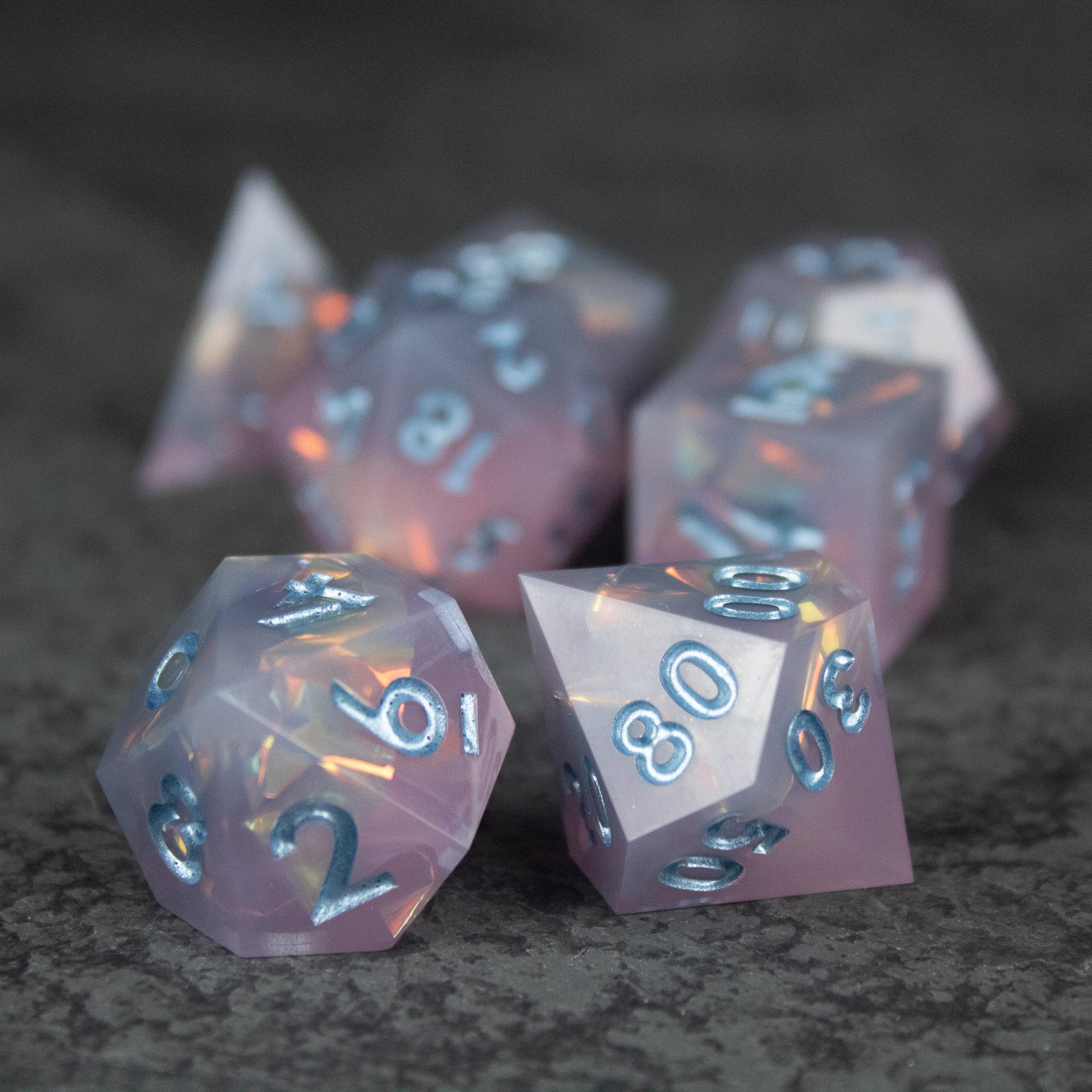 Moonshard Dice Set - Translucent pink sharp-edged polyhedral dice set with blue numbers on a dark surface.
