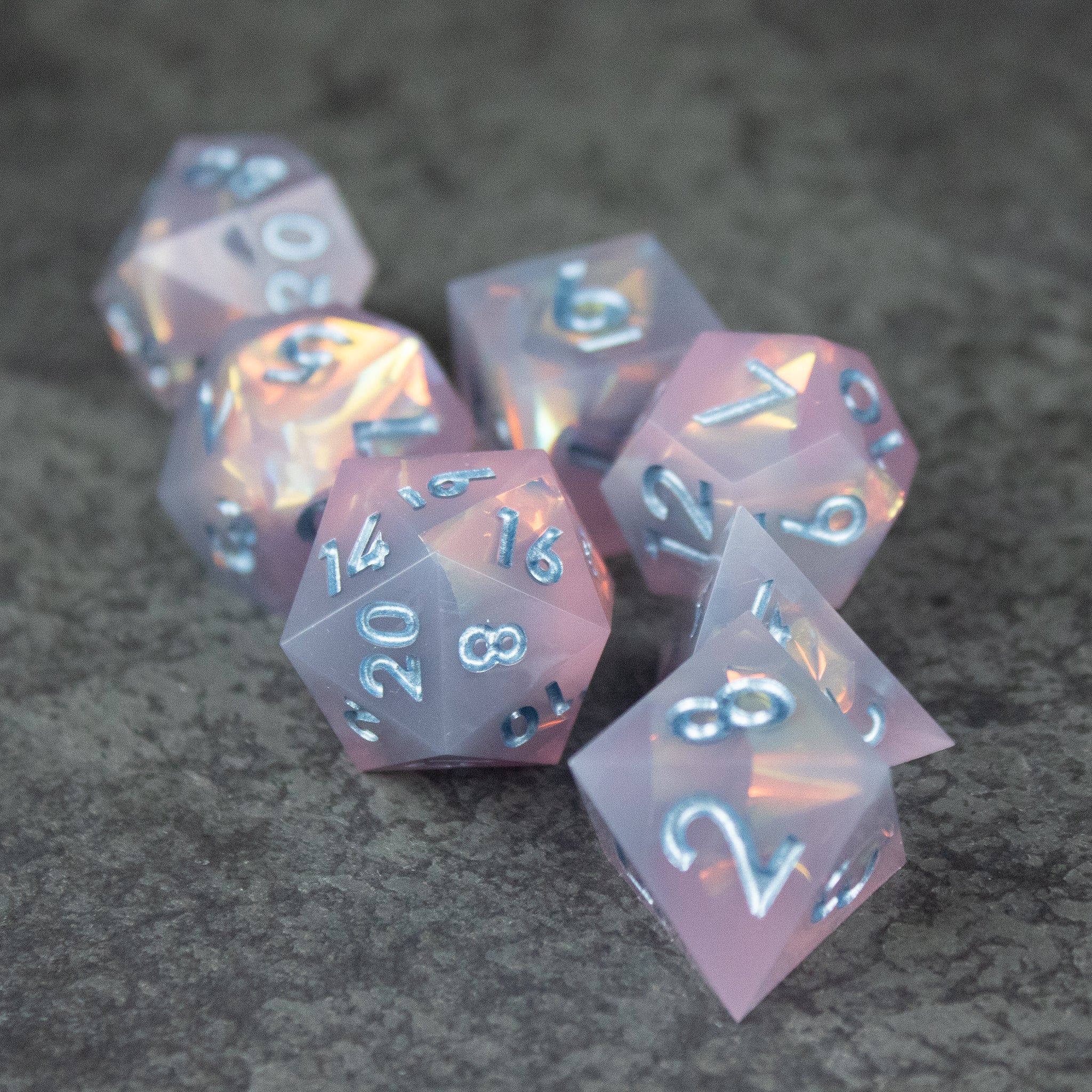 Moonshard Dice Set - Translucent pink sharp-edged polyhedral dice set with white numbering on textured gray surface.