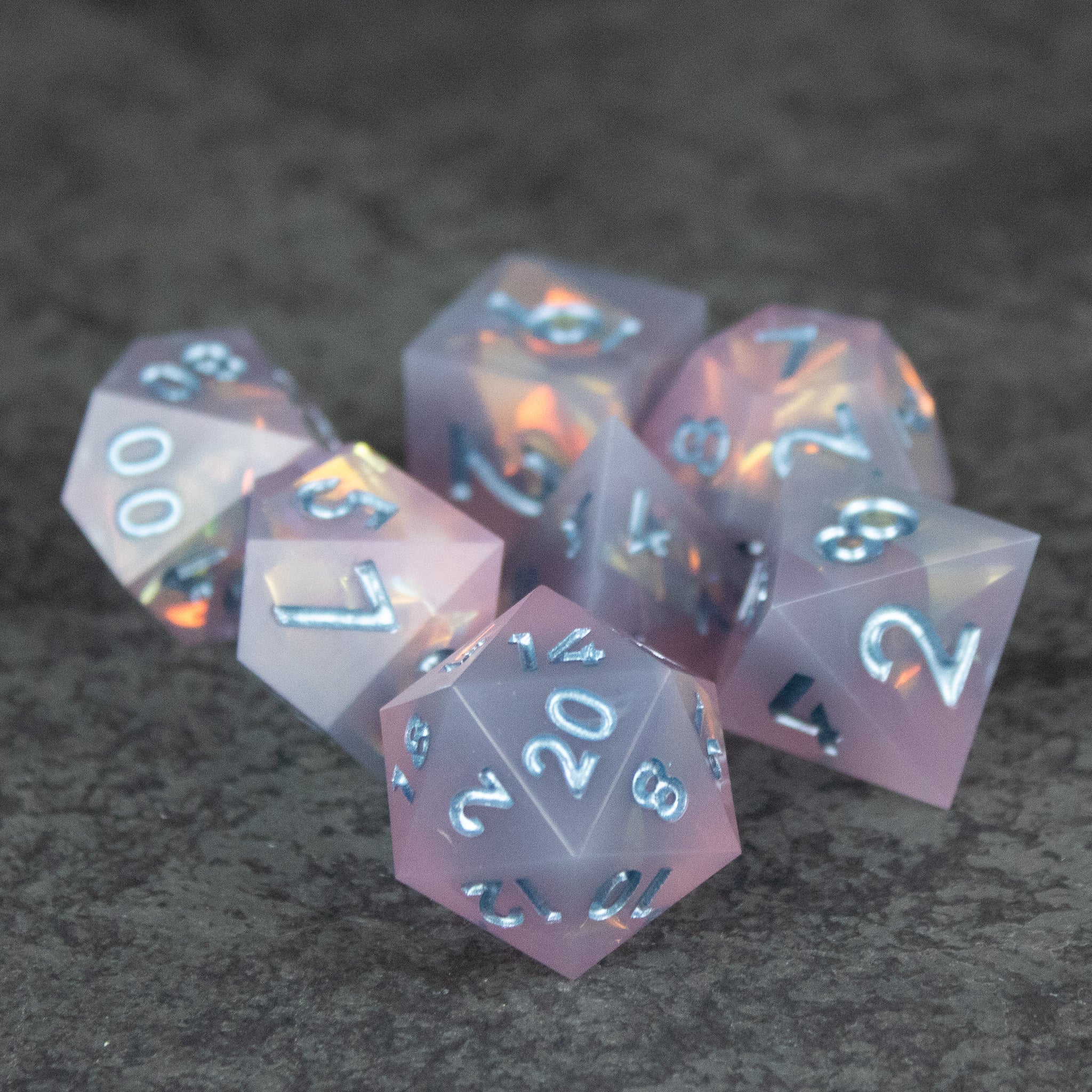 Moonshard Dice Set - Set of translucent pink polyhedral dice with sharp edges and white numbering on a dark textured surface.