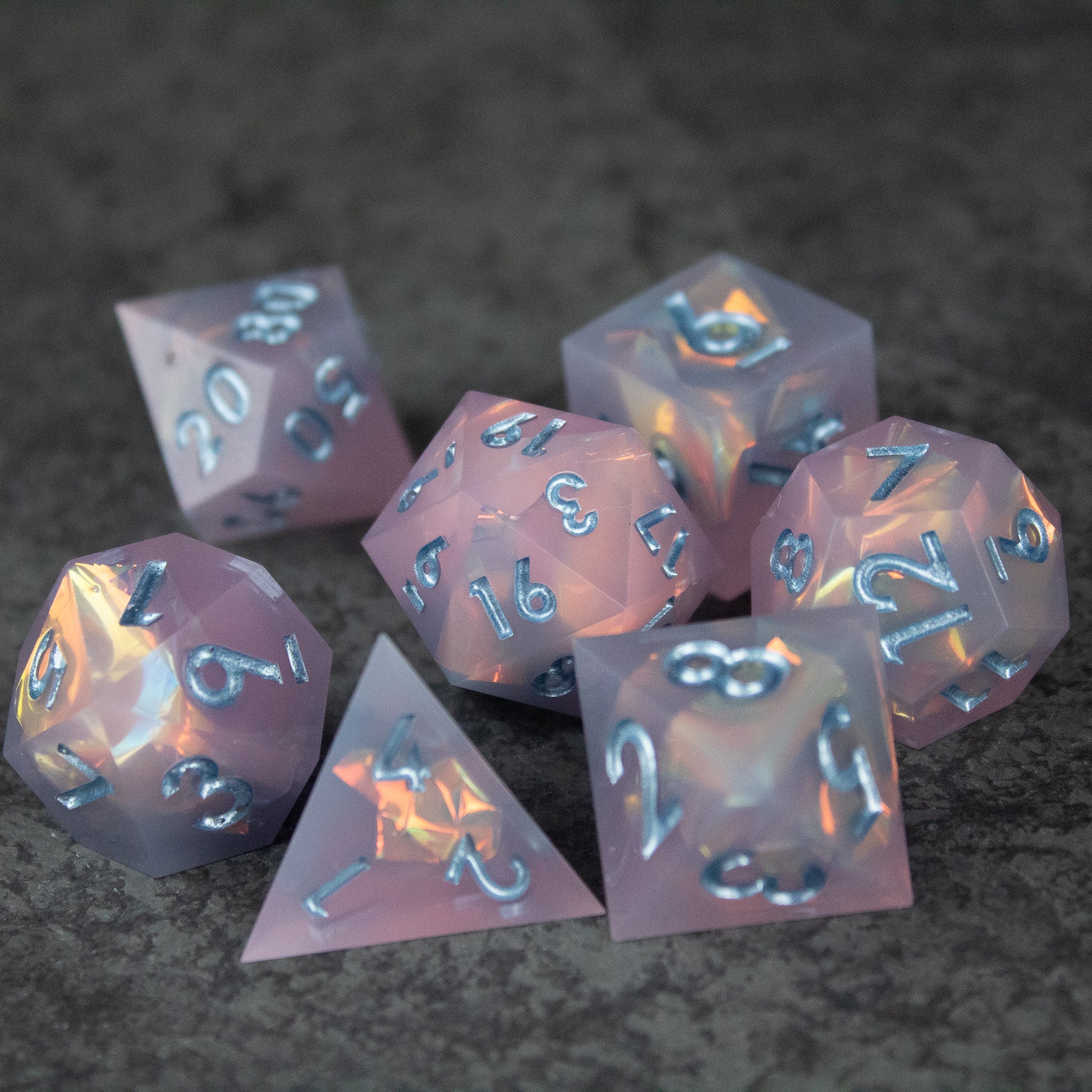 Moonshard Dice Set - Set of seven translucent pink sharp-edged polyhedral dice with metallic silver numbers on a dark textured surface.