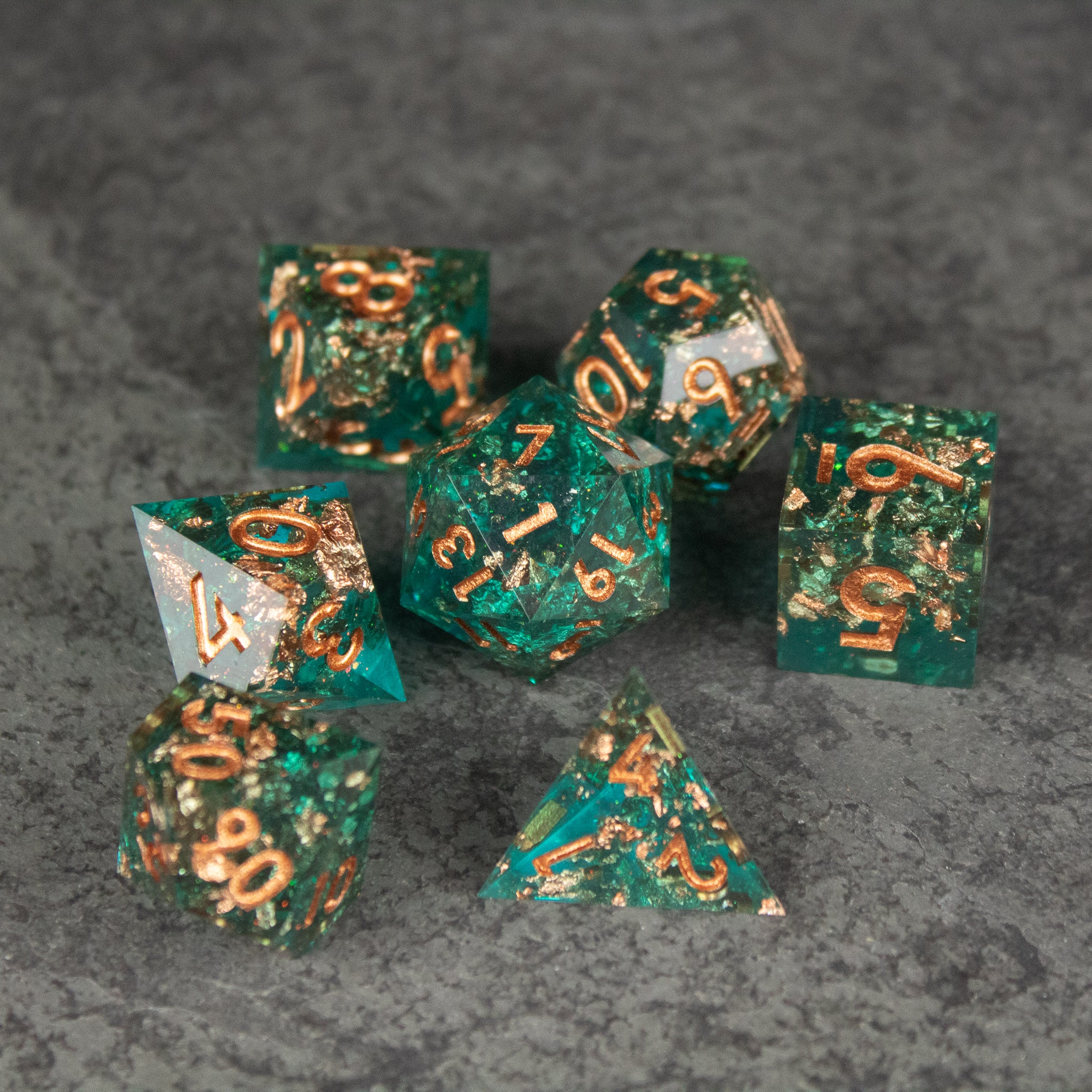 Oasis Dice Set - Set of seven translucent emerald green polyhedral dice with gold numbering and sharp edges on a gray textured surface.