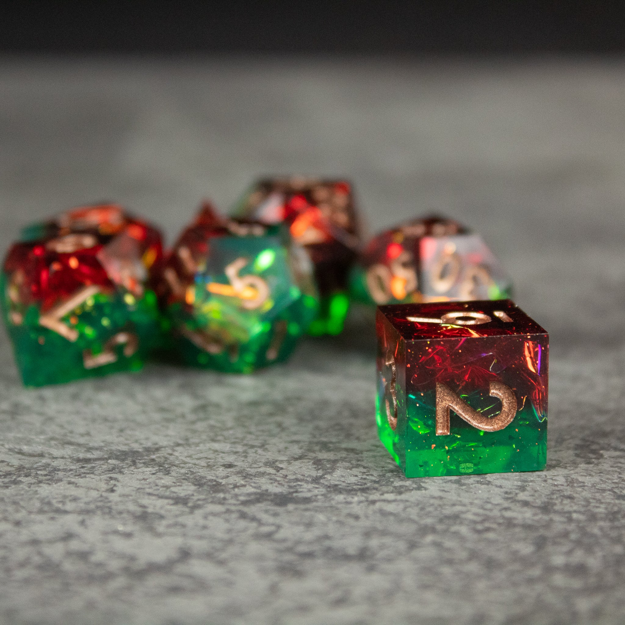Jungle Fire Dice Set - Sharp-edged translucent dice with red and green gradient and gold numbers on a gray surface.