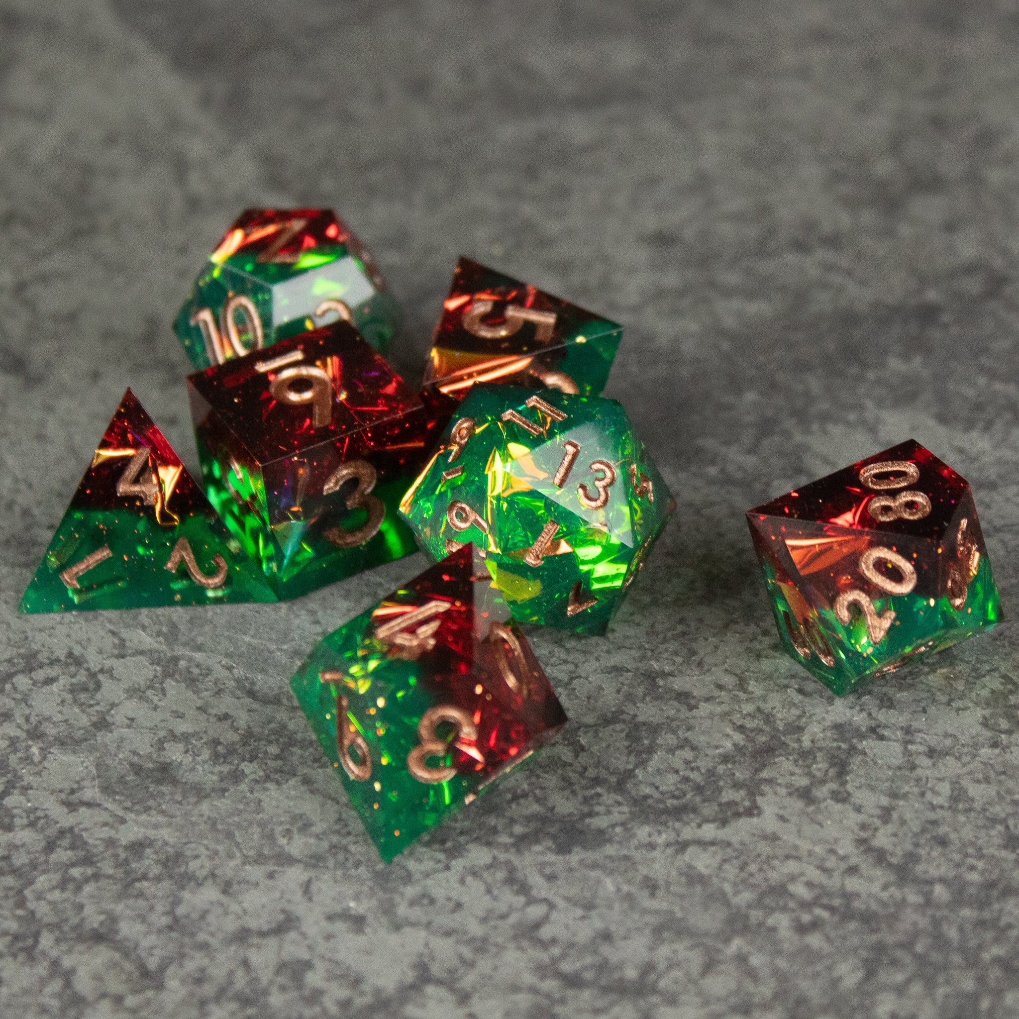 Jungle Fire Dice Set - Seven sharp-edged green and red translucent polyhedral dice with gold numbers on gray textured surface.