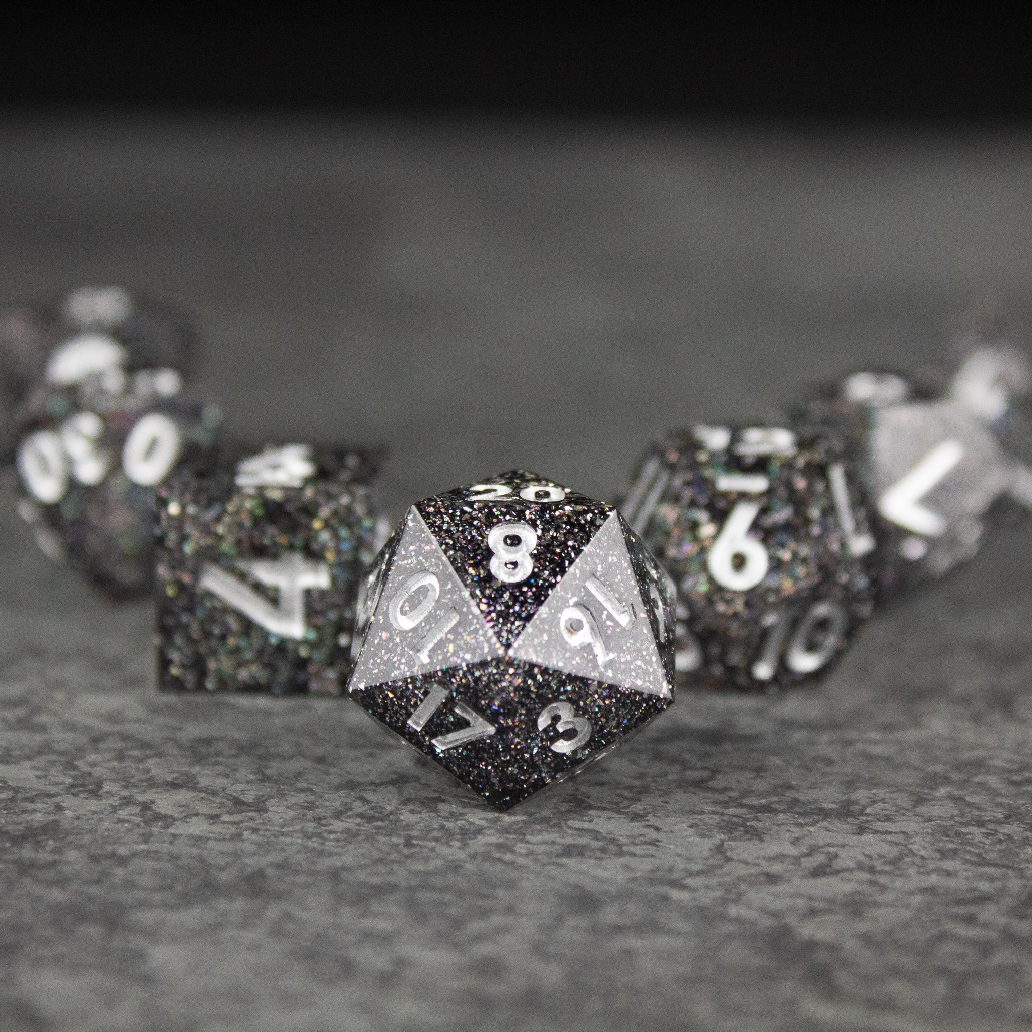 Starfield Dice Set - Black and white speckled 20-sided die with sharp edges and white numbering, surrounded by similar dice on gray surface.