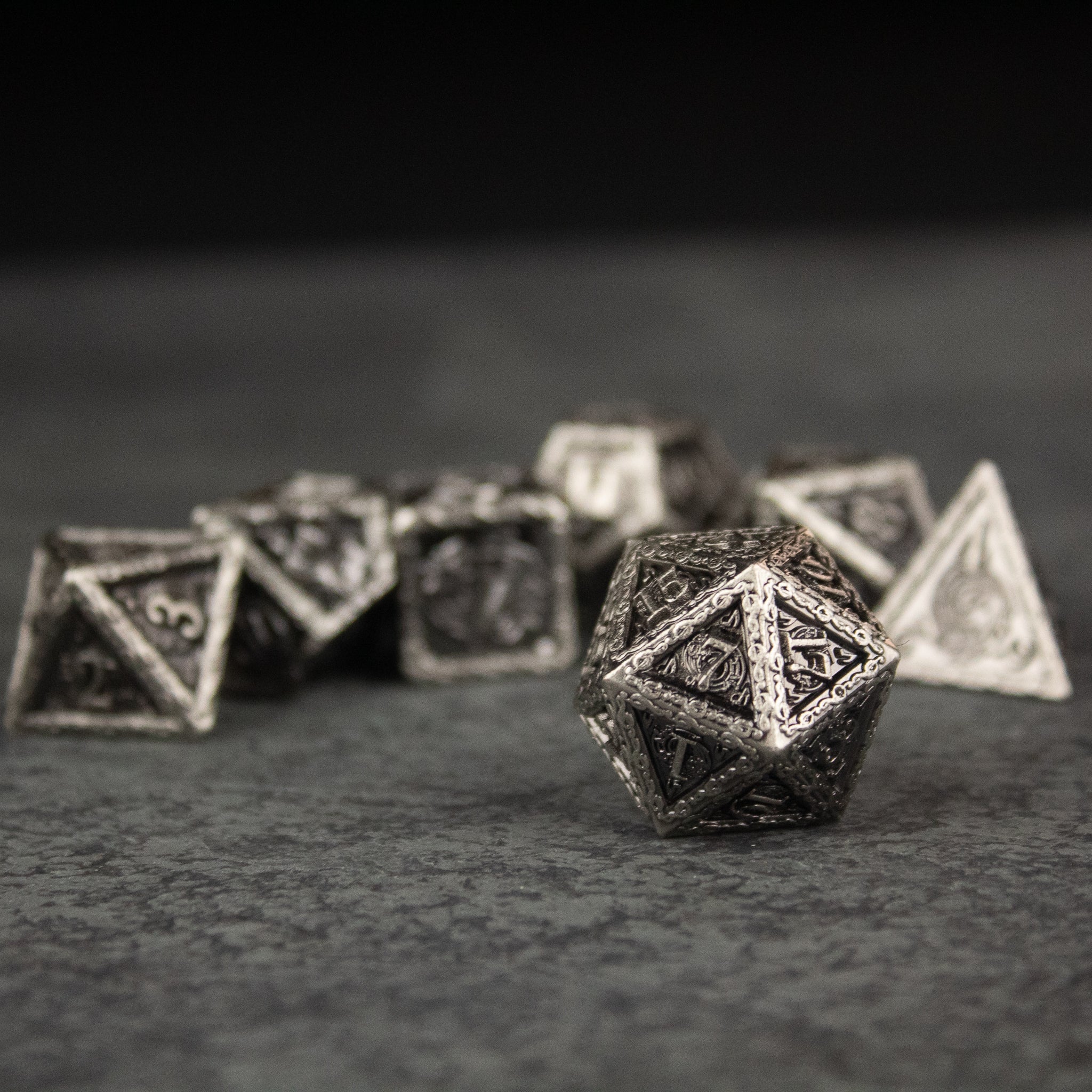 Bardic Inspiration Metal Dice Set - Metallic silver polyhedral dice set with intricate runic designs, featuring a d20 in focus and other dice blurred in the.