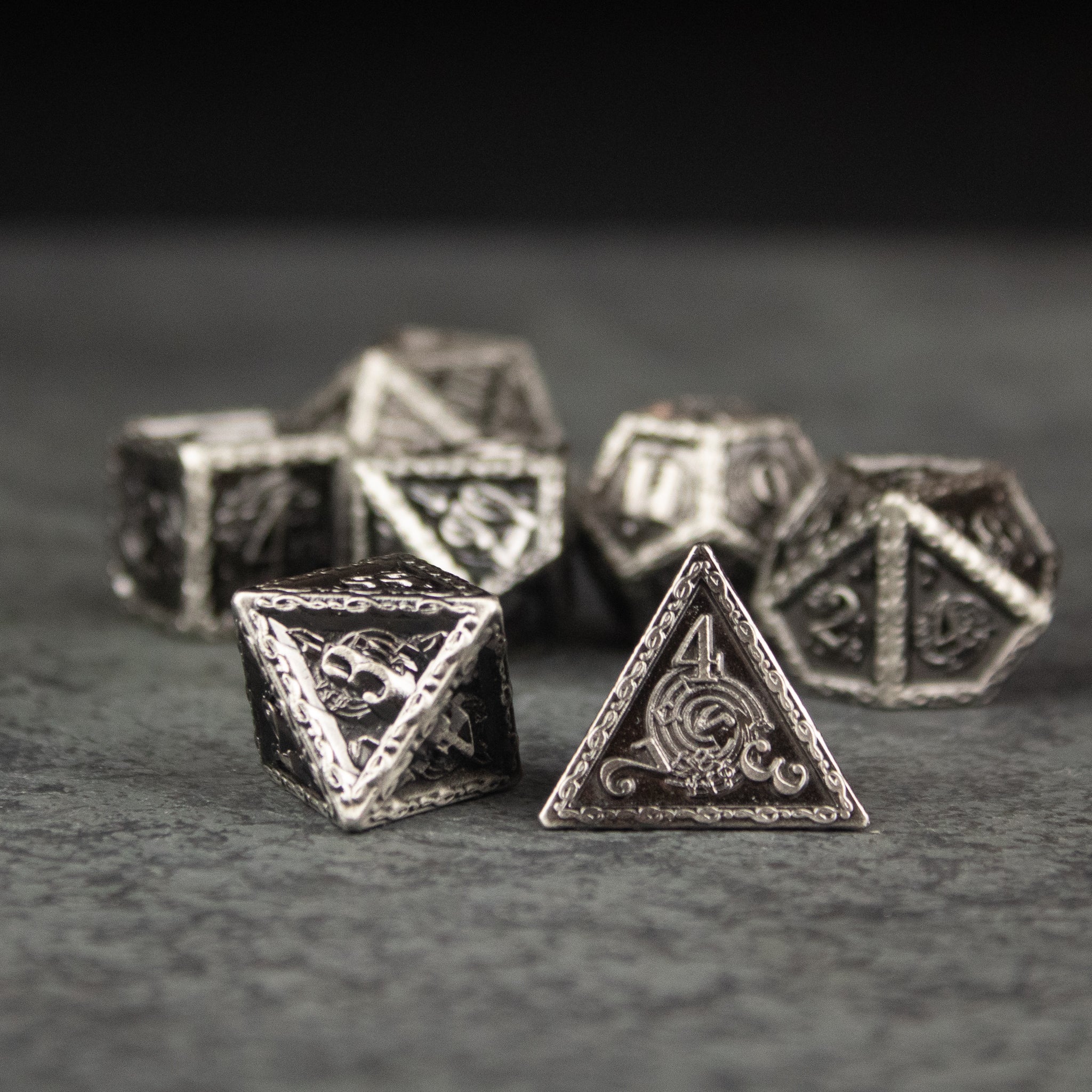 Bardic Inspiration Metal Dice Set - Antique silver metal dice set with intricate engravings and decorative symbols, including a d4 in front.