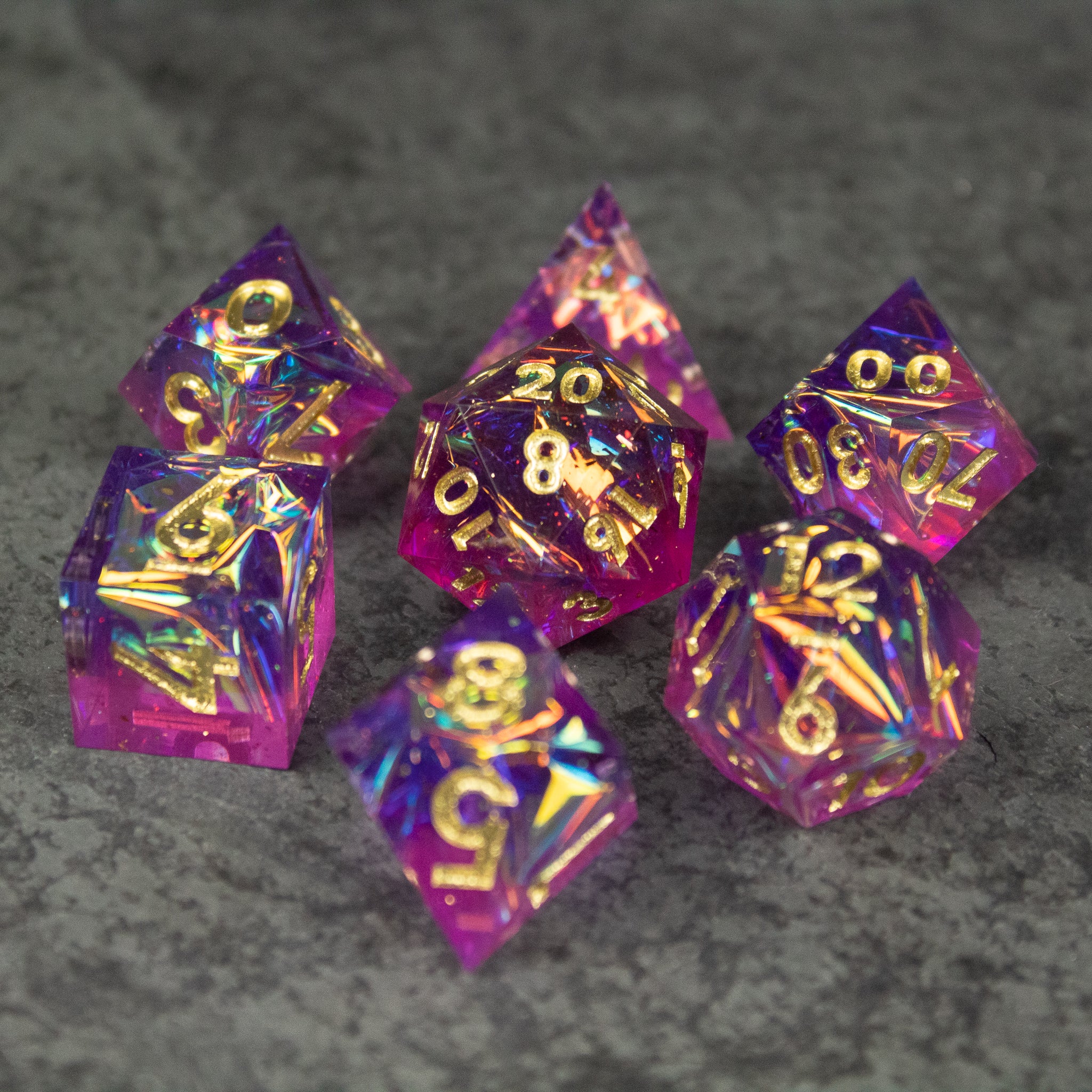 Aether Dice Set - Set of seven translucent pink and purple sharp-edged polyhedral dice with gold numbers on gray surface.