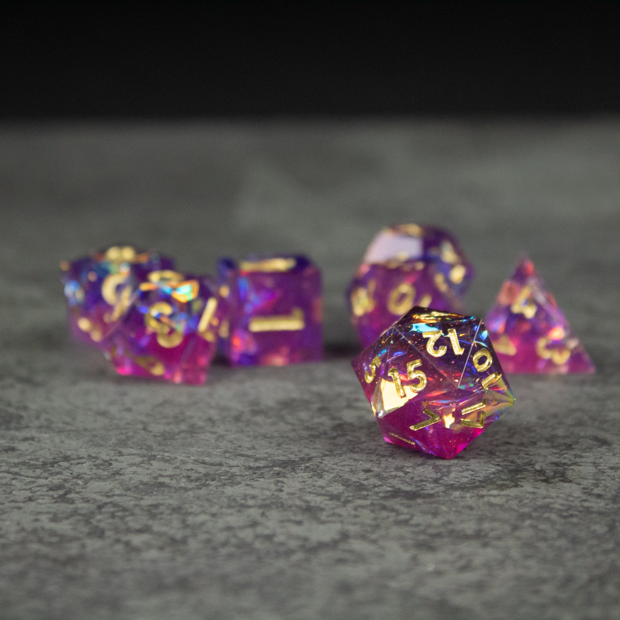 Aether Dice Set - Seven sharp-edged polyhedral dice in translucent pink and purple with gold numbers on a gray surface.