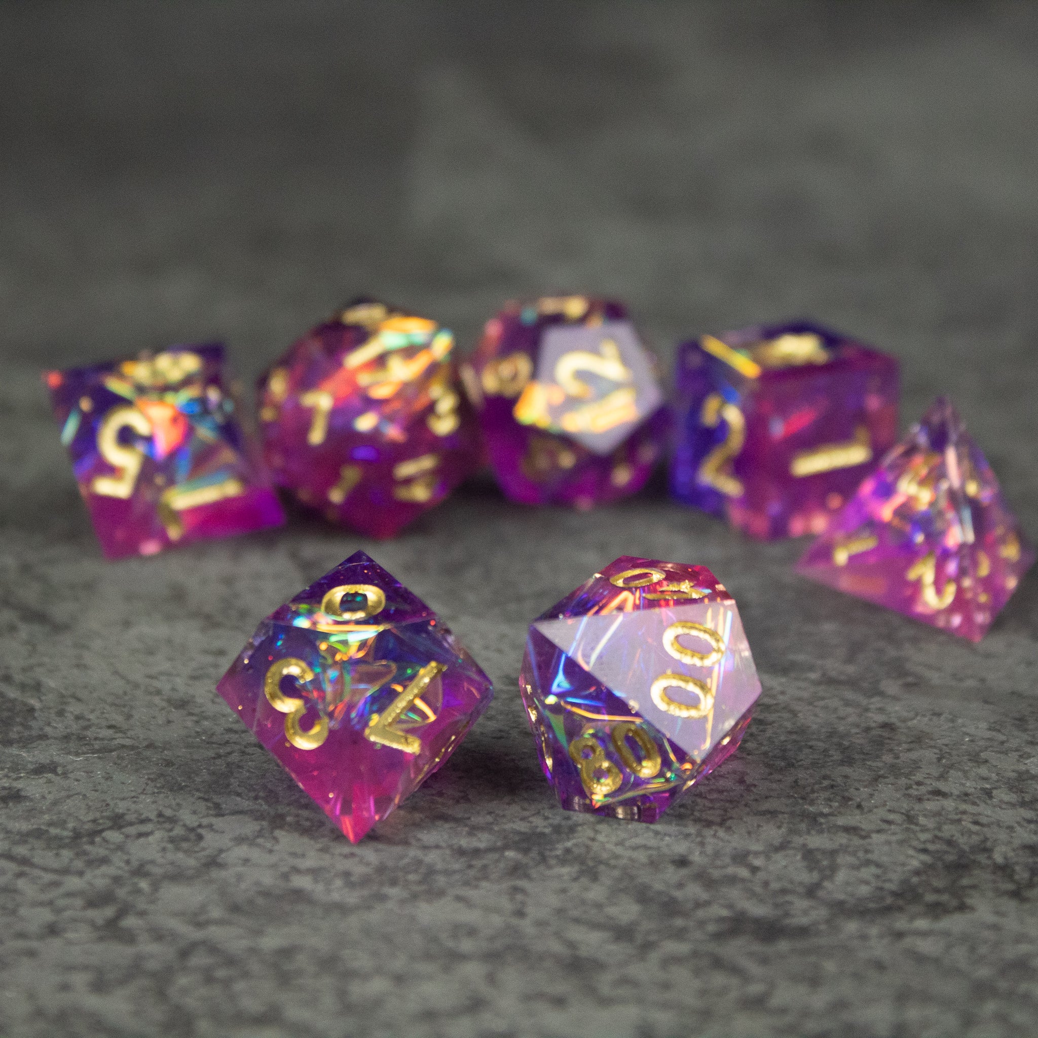 Aether Dice Set - Set of seven sharp-edged polyhedral dice in translucent pink and purple with gold flecks and white numbering.