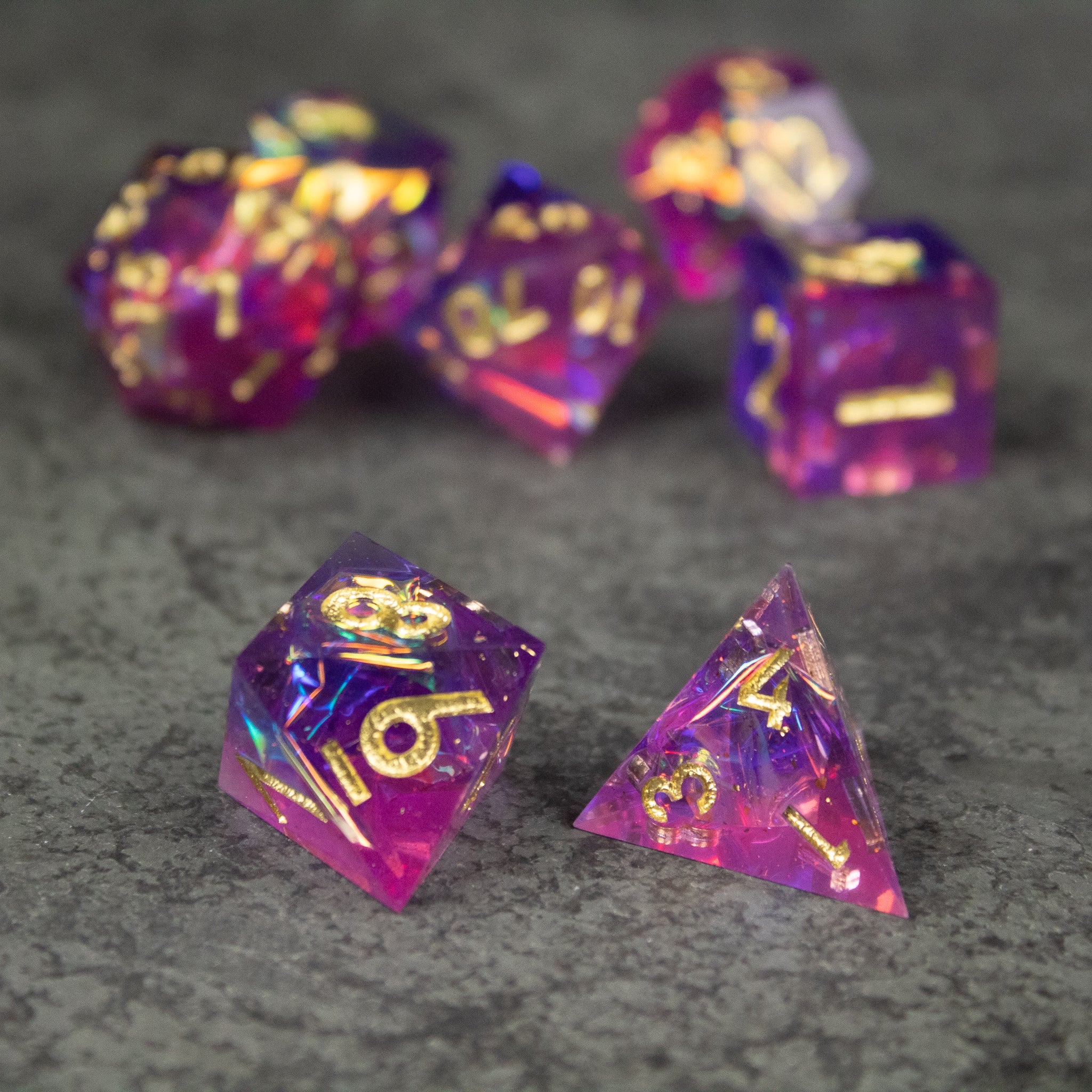 Aether Dice Set - Set of translucent sharp-edged dice in pink and purple hues with gold numbers on a gray surface.