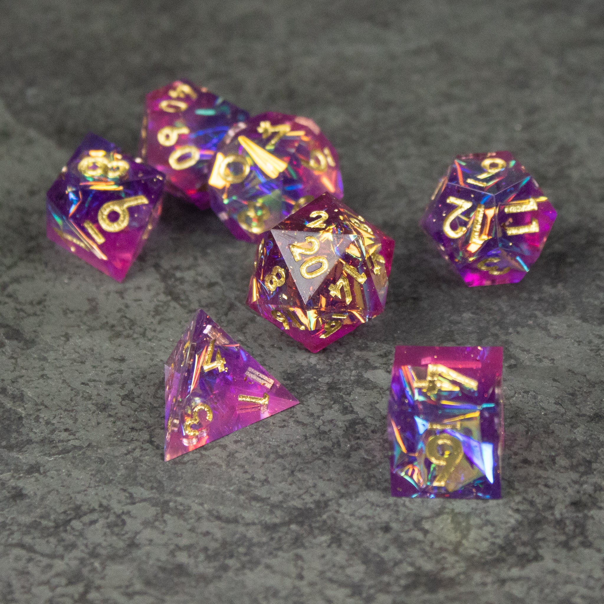Aether Dice Set - Seven sharp-edged dice in pink and purple translucent resin with gold numbers on gray textured surface.
