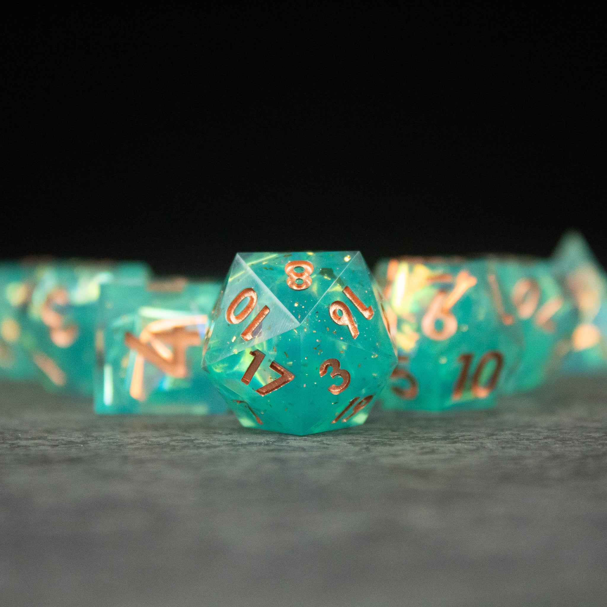 Residuum Dice Set - Transparent aquamarine 20-sided dice with sharp edges and bronze numbers in focus with matching dice blurred in background.