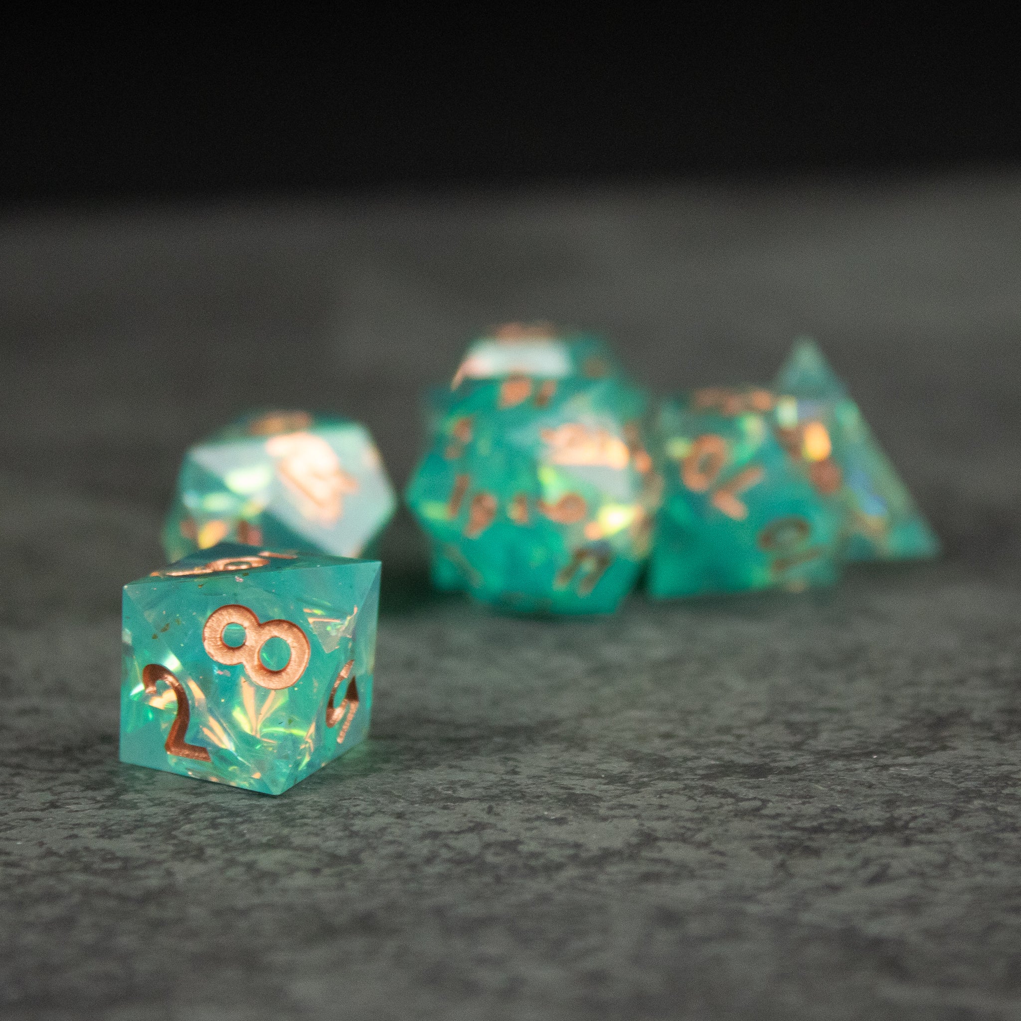 Residuum Dice Set - Translucent aquamarine sharp-edged dice set with gold numbering on gray surface.