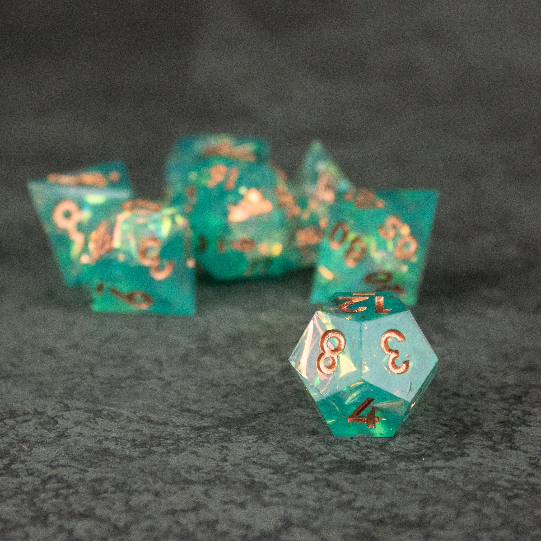 Residuum Dice Set - Translucent aquamarine sharp-edged dice set with metallic gold numbers on a dark gray surface.