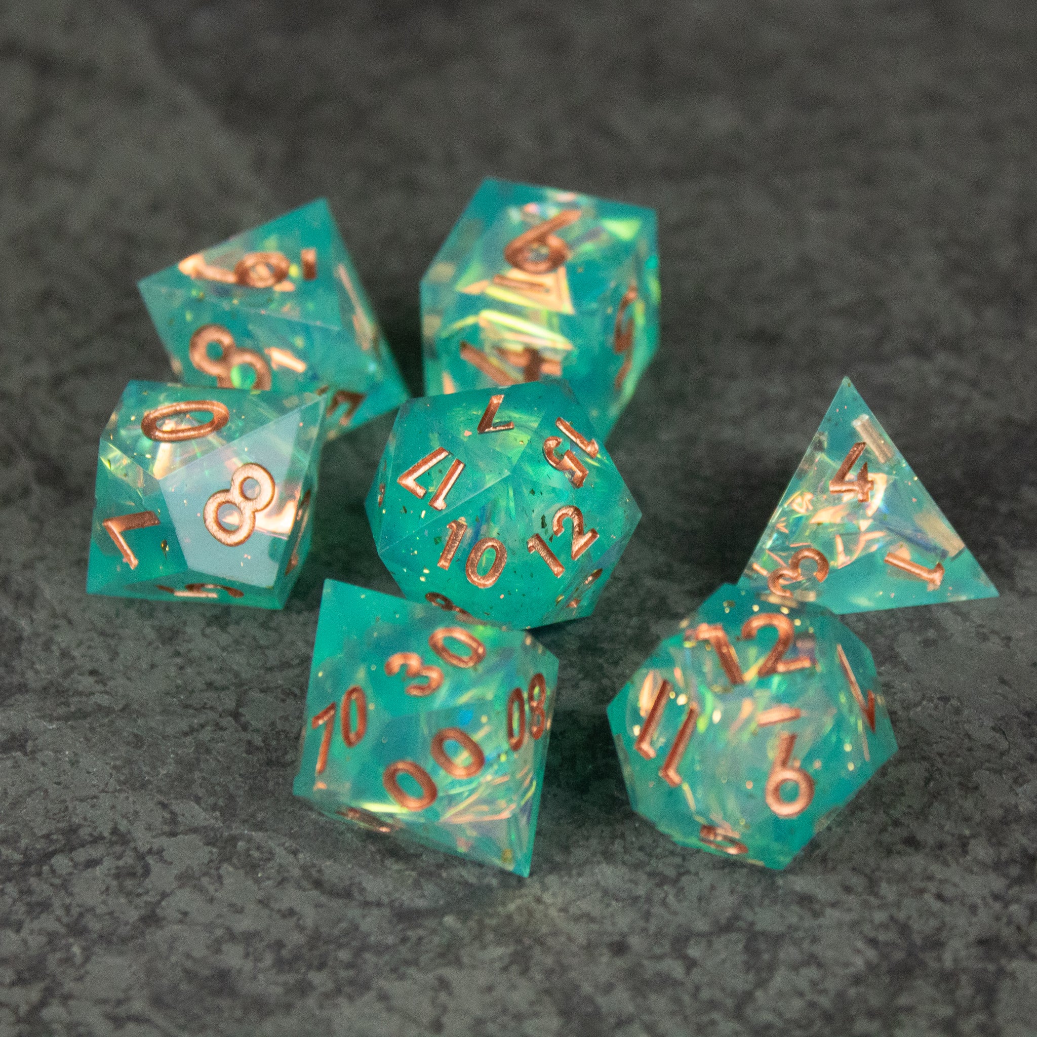 Residuum Dice Set - Set of seven transparent aquamarine sharp-edged dice with gold numbers on a dark gray surface.