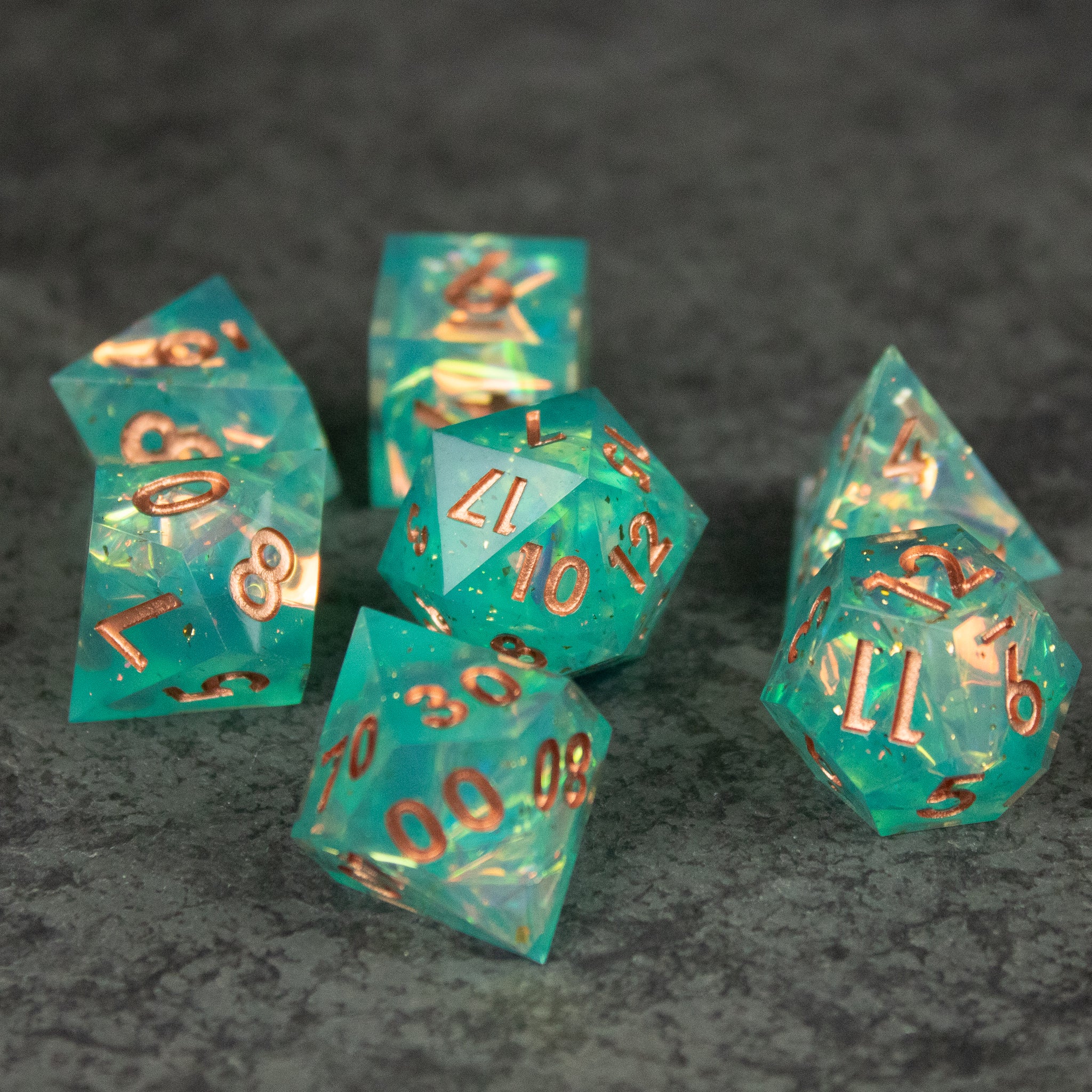 Residuum Dice Set - Set of seven aquamarine translucent sharp-edged dice with gold numbering on dark textured surface.