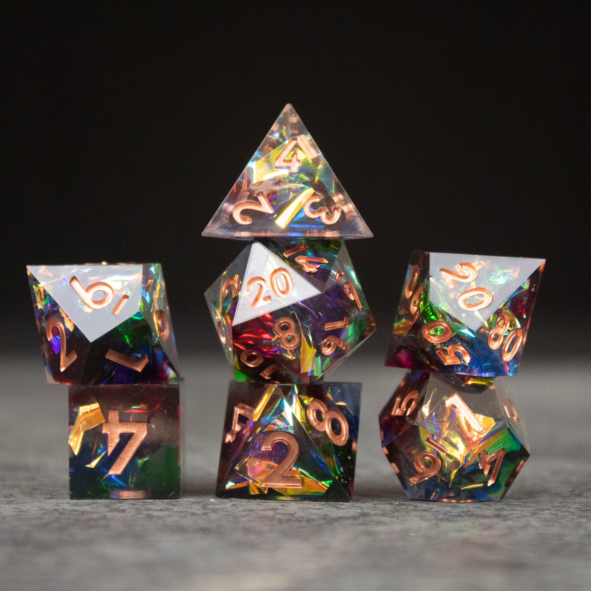 Aurora Dice Set - Set of seven sharp-edged rainbow dice with iridescent colors and gold numbers on a gray surface.