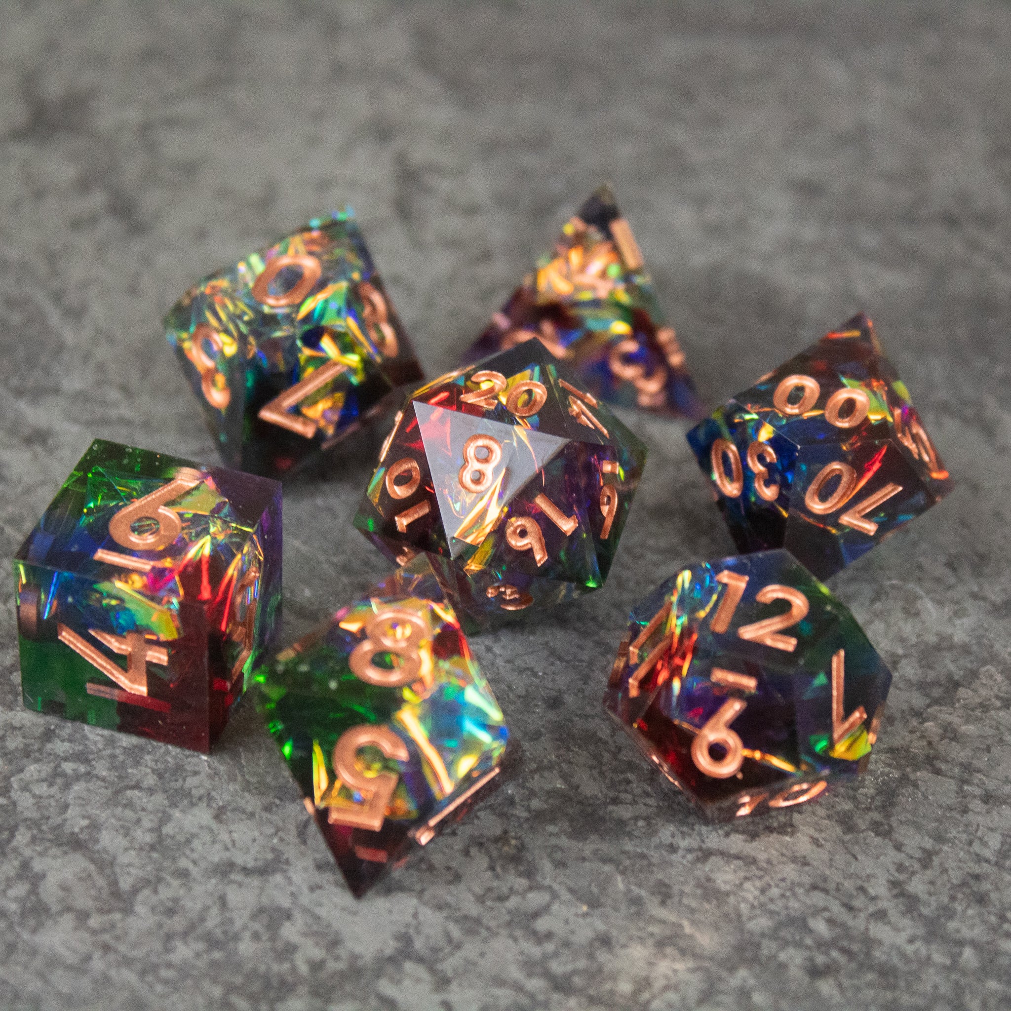 Aurora Dice Set - Set of sharp-edged rainbow polyhedral dice with gold numbers on gray stone surface.