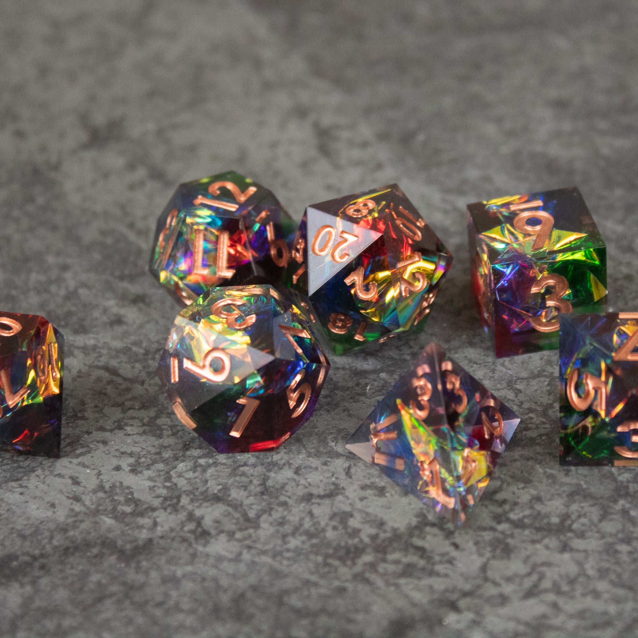 Aurora Dice Set - Set of transparent, rainbow-colored sharp edge dice with gold numbers on a gray textured surface.
