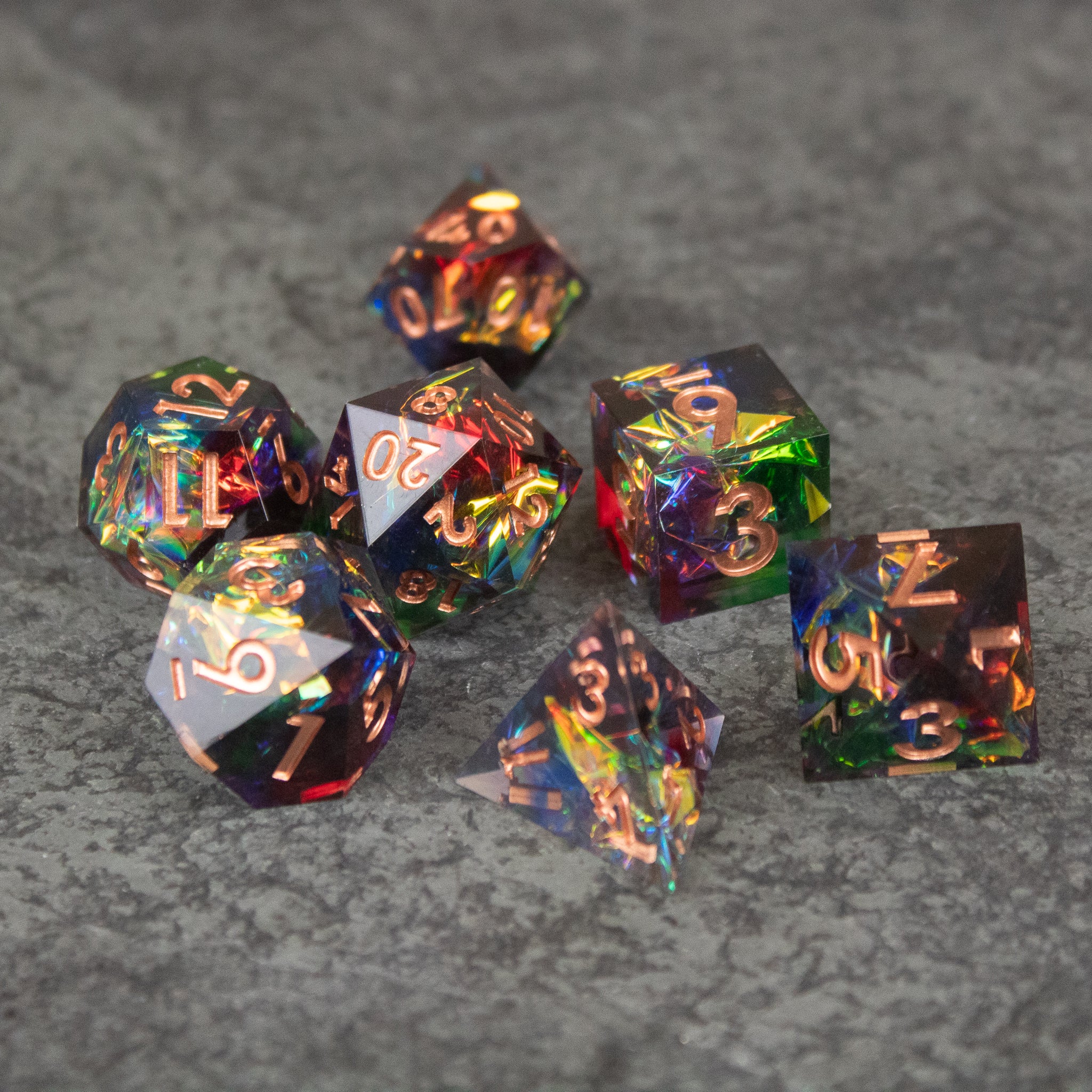 Aurora Dice Set - Seven sharp-edged translucent dice with rainbow colors and gold numbers on a textured gray surface.
