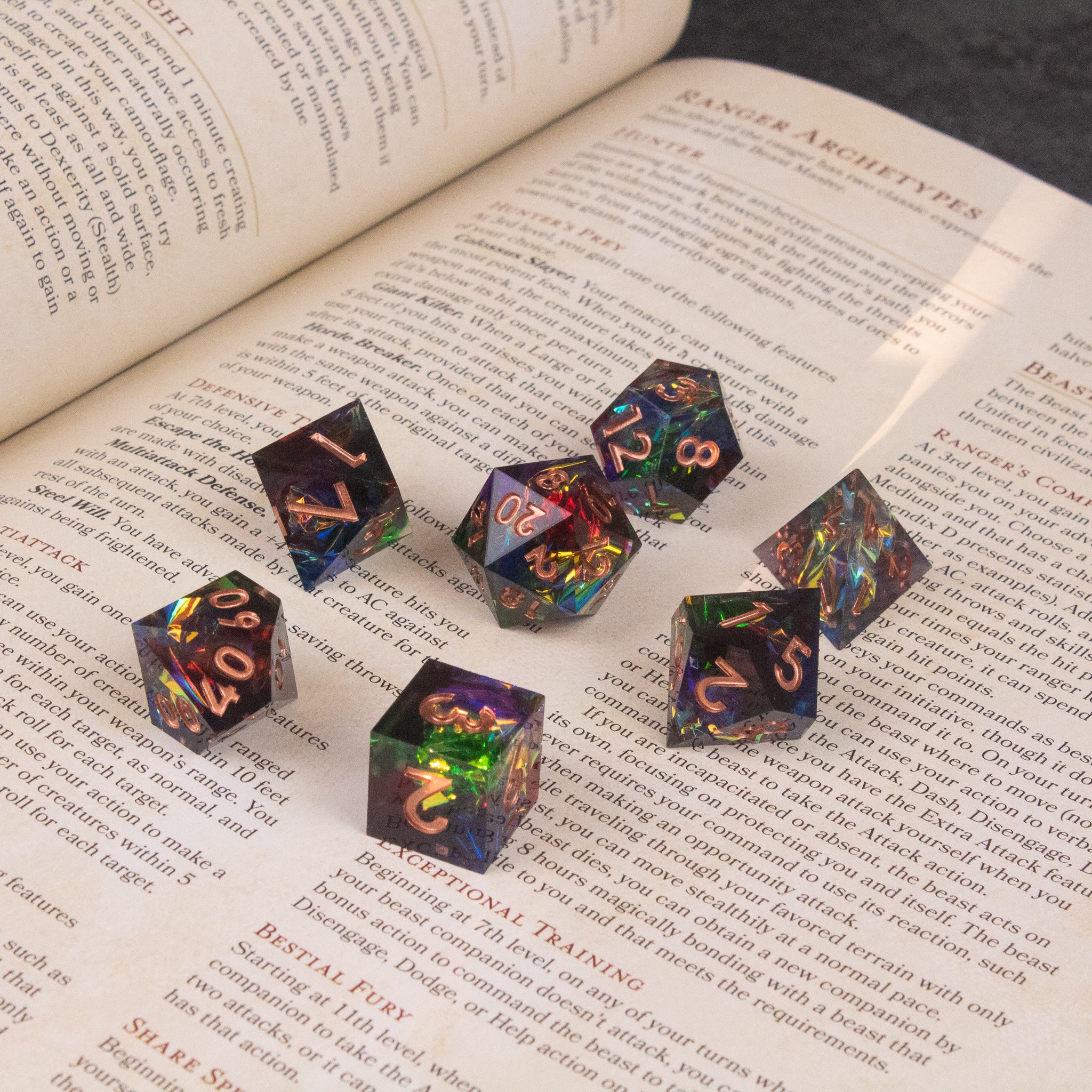 Aurora Dice Set - Seven sharp-edged polyhedral dice set with translucent rainbow colors and gold numbers on an open book.