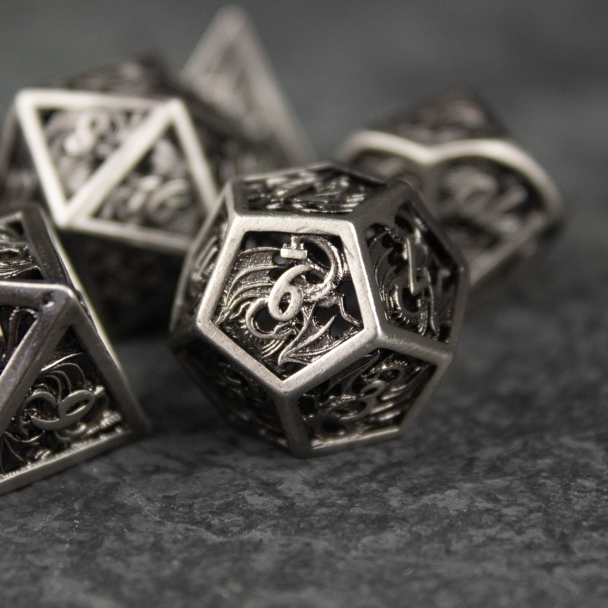 Dragon Cult Hollow Dice Set - Metallic hollow polyhedral dice set with intricate dragon-themed cutouts and etched numbers in silver-gray finish.