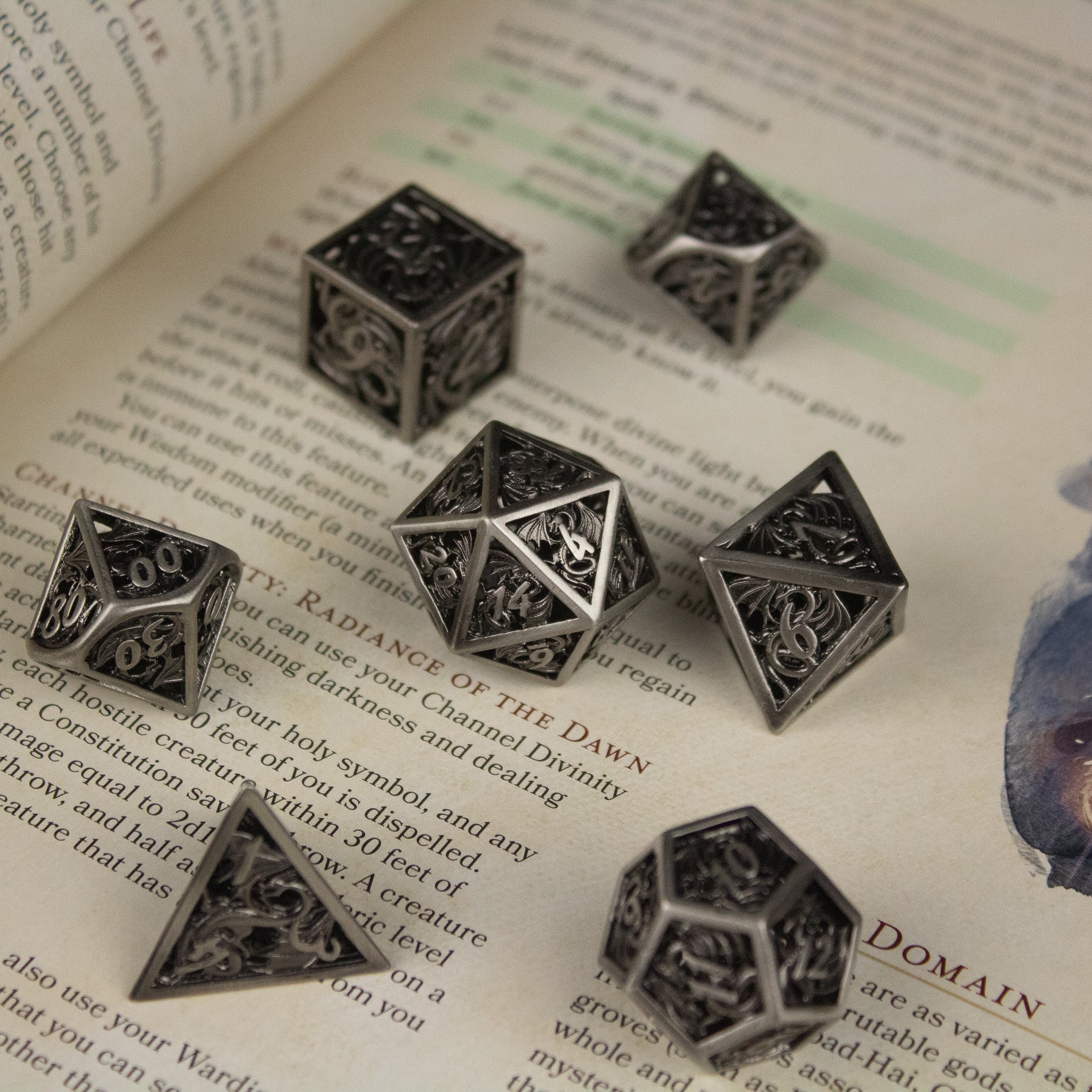 Dragon Cult Hollow Dice Set - Set of six black metal hollow dice with intricate dragon engravings and silver numbering, displayed on an open book page.