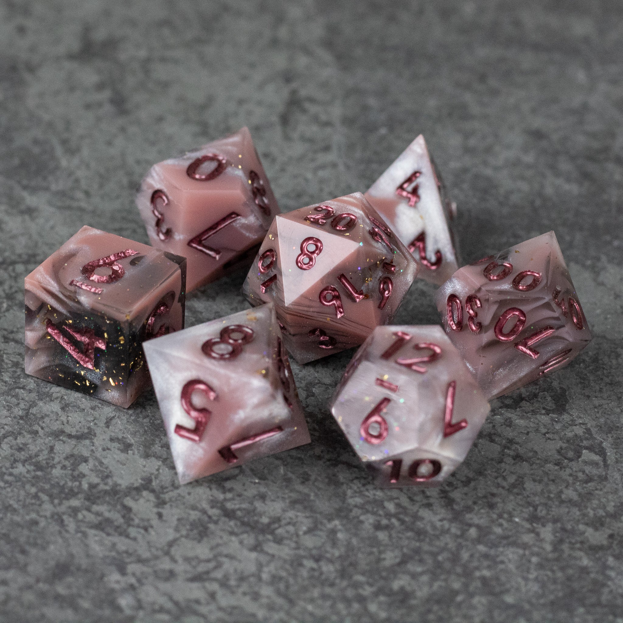 Rosestone Dice Set - Set of seven red and white marbled sharp edge dice with gold and red numbering on gray textured surface.