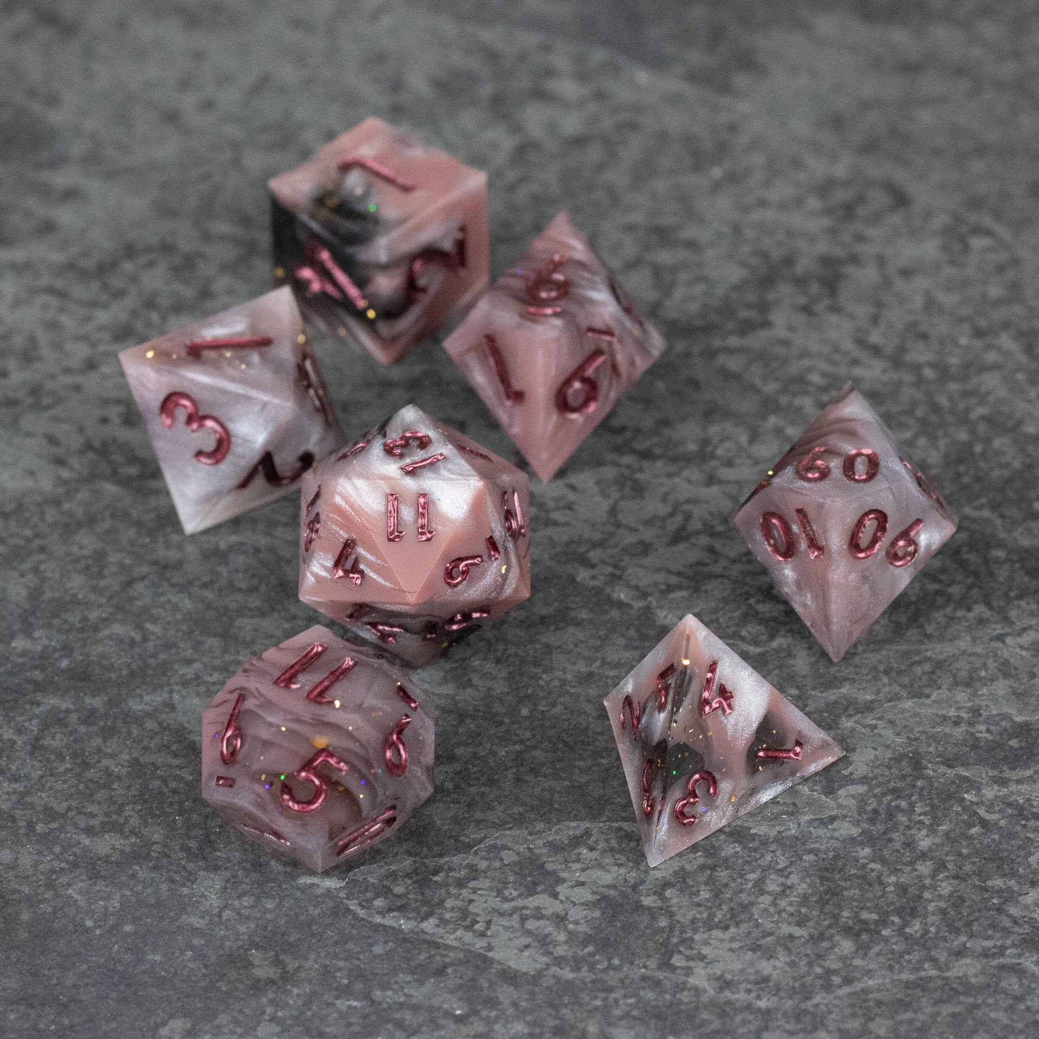 Rosestone Dice Set - Set of seven translucent red sharp-edged dice with rose gold flecks on a gray surface.