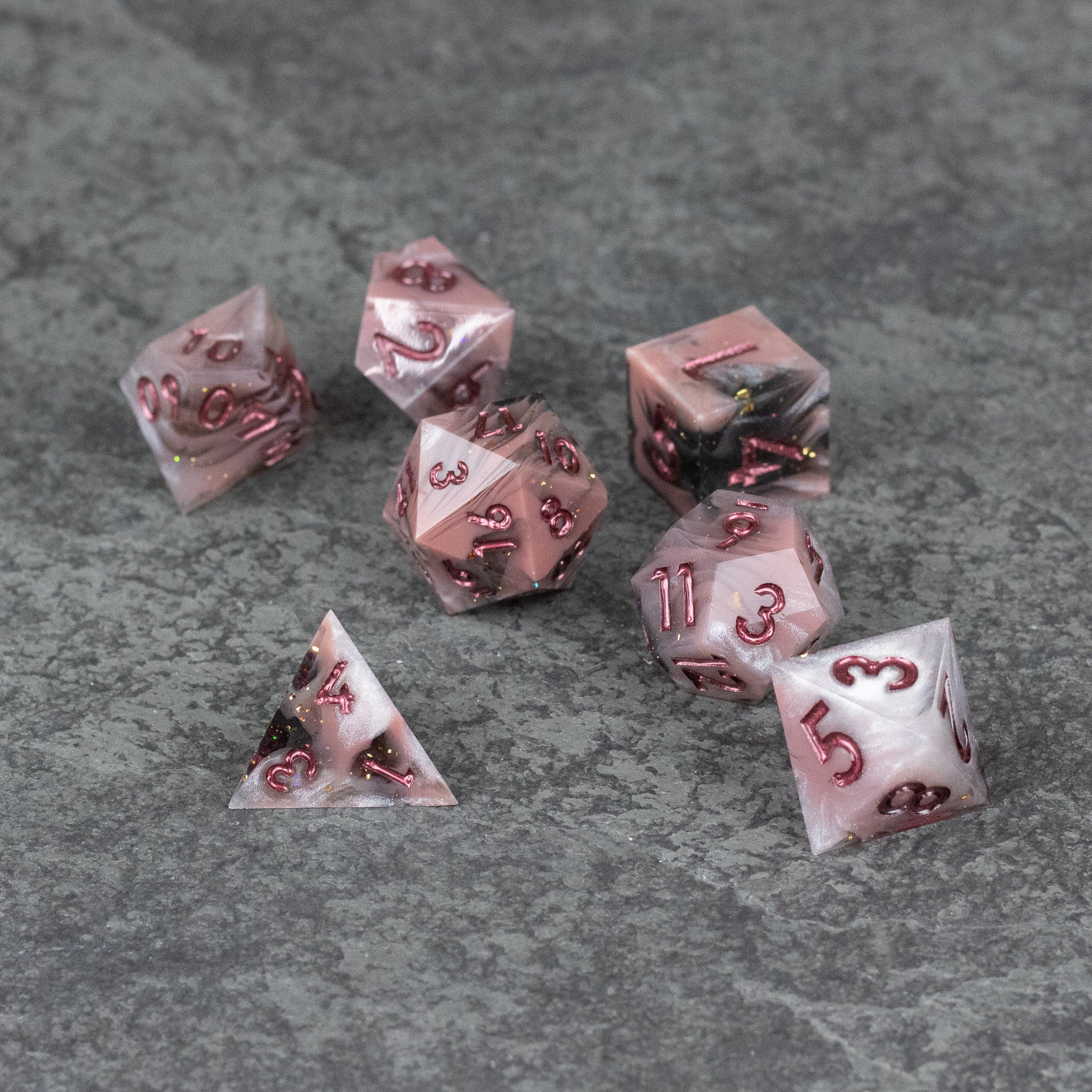 Rosestone Dice Set - Seven translucent red sharp-edged polyhedral dice with metallic numbers on gray textured surface.