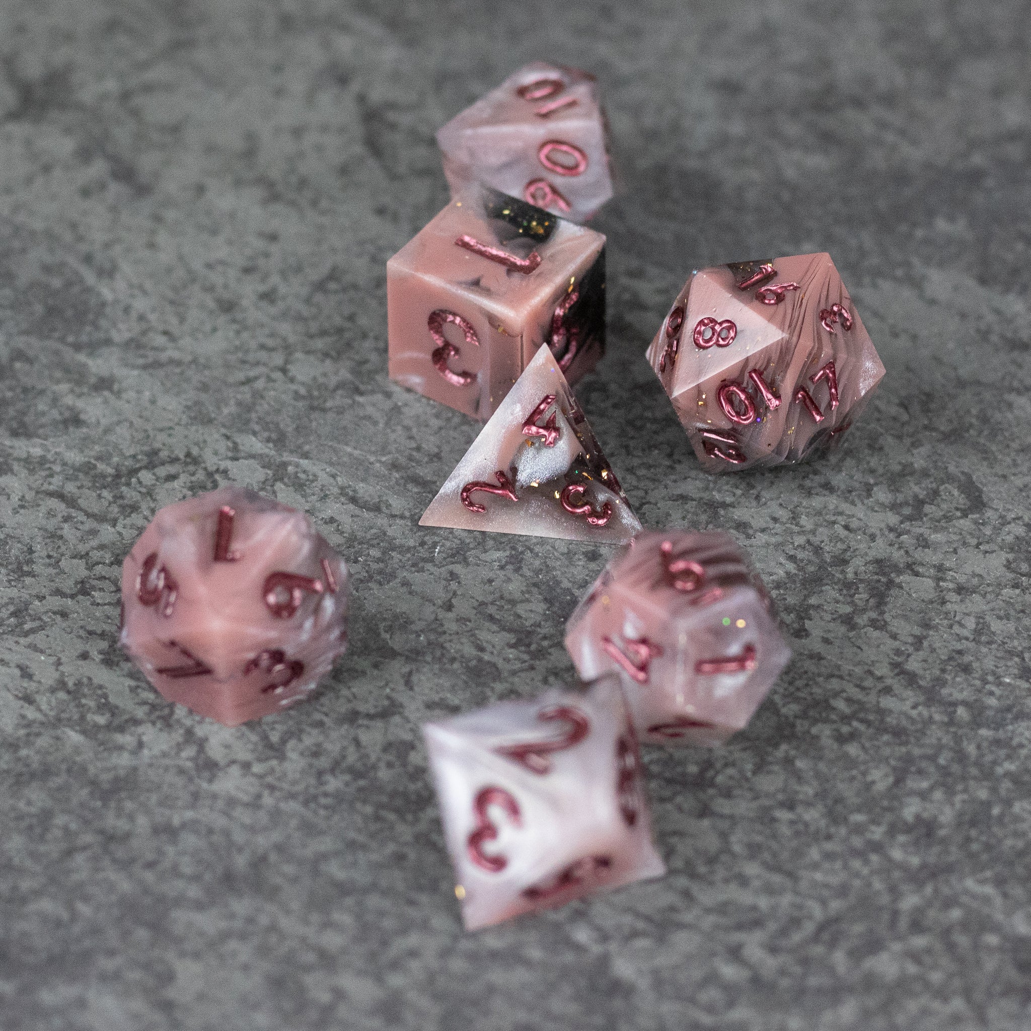 Rosestone Dice Set - Seven translucent pink polyhedral dice with sharp edges and red numbering on gray textured surface.