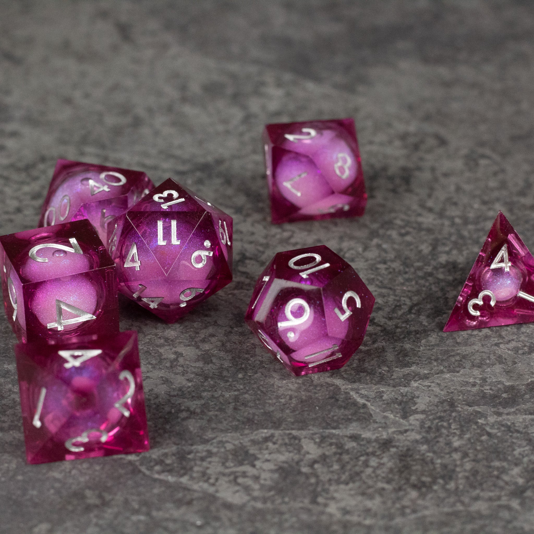 Magic Wine - Liquid Core Dice Set - Set of seven sharp-edged, translucent red polyhedral dice with swirling liquid core and white numbers on a gray textured.
