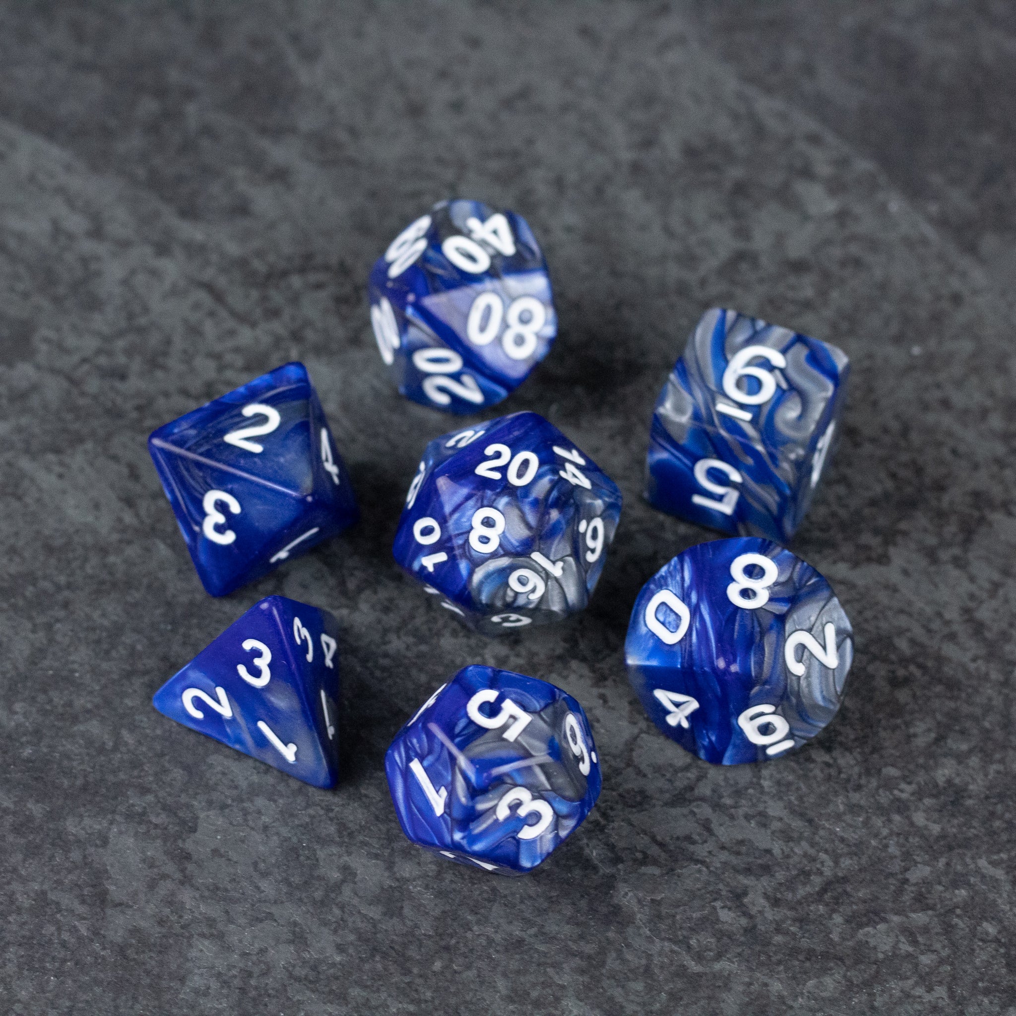 Royal Blue and Silver Acrylic Dice Set - Seven royal blue acrylic dice with white numbering, including d20, d12, d10, d8, d6, and d4, resting on dark textured.