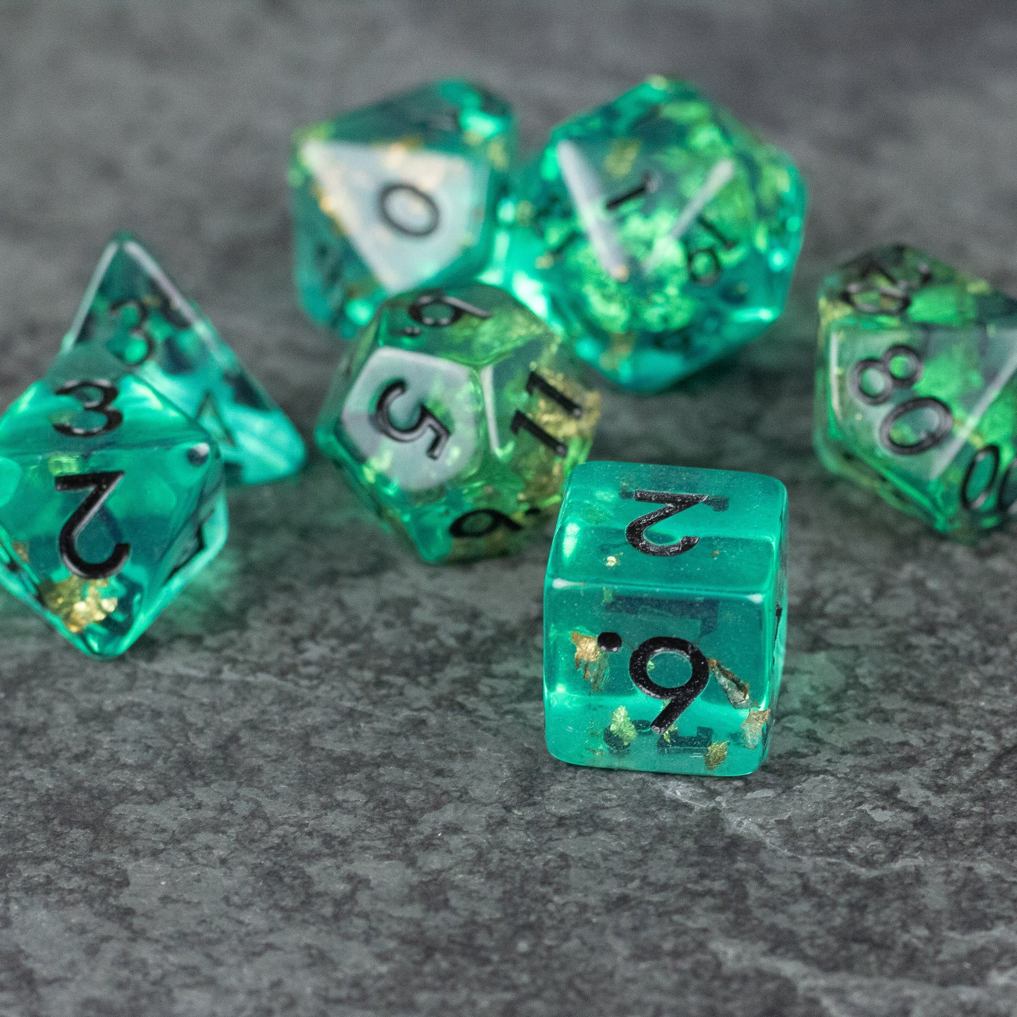 Teal Foil Dice Set - Set of seven teal translucent polyhedral dice with gold foil flecks and black numbers on a gray textured surface.