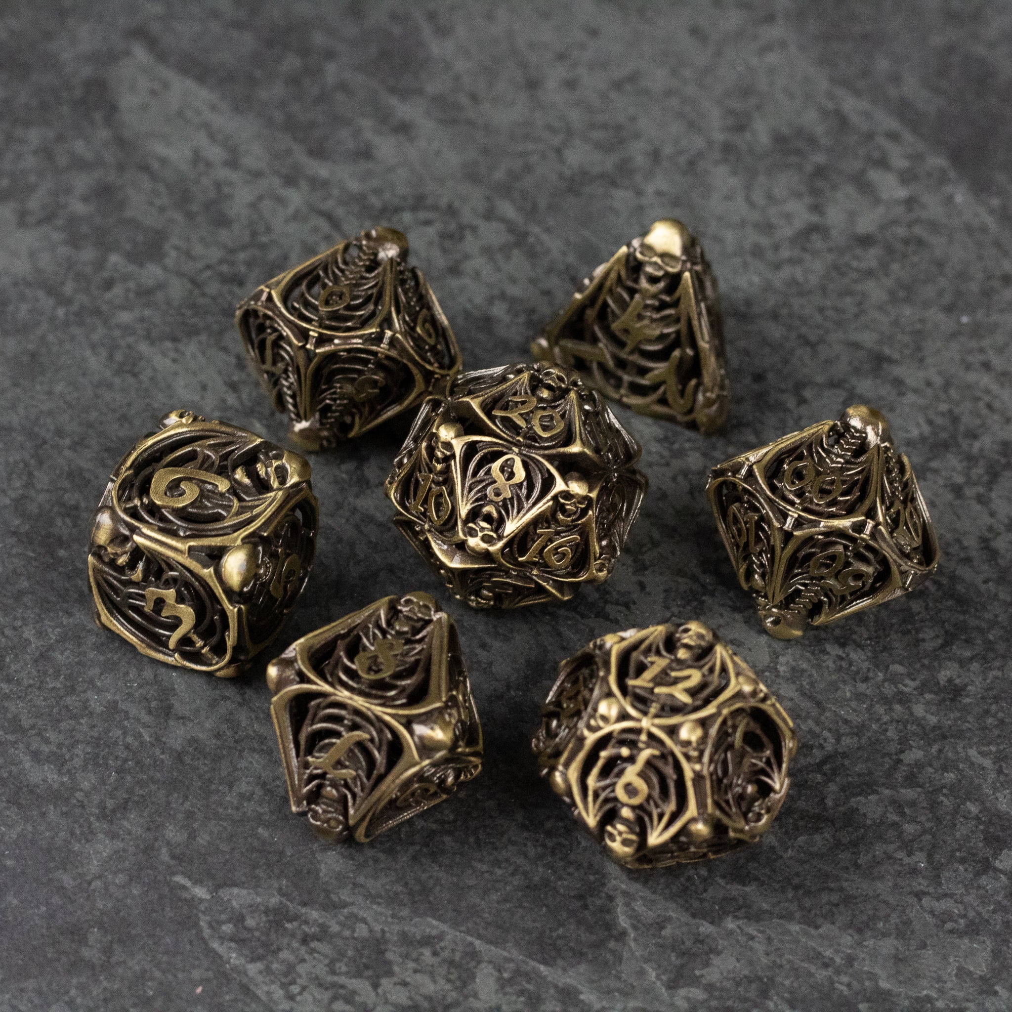 Bone Crypt Dice Set - Metallic gold hollow dice set with intricate carved bone-like patterns, seven pieces on gray surface.
