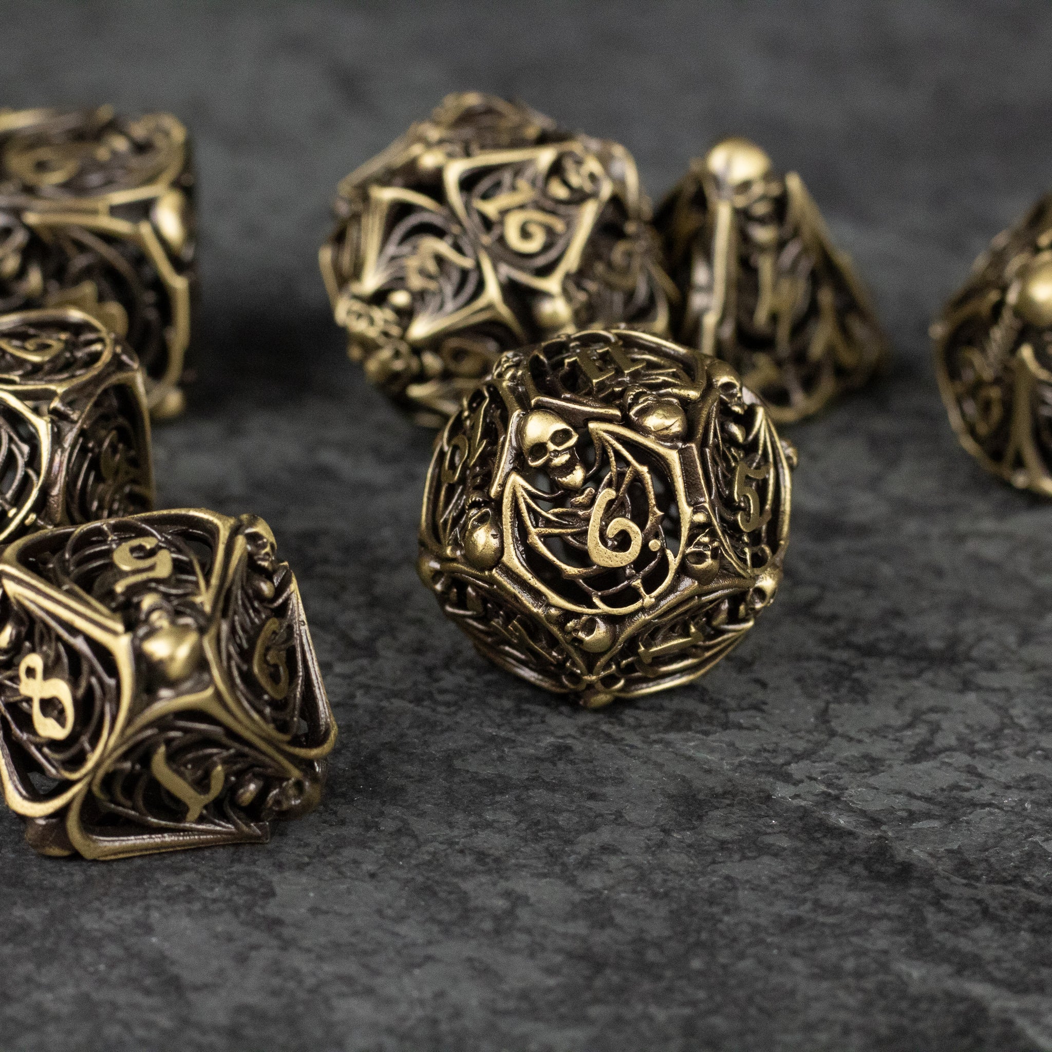 Bone Crypt Dice Set - Antique gold metal dice set with intricate hollow designs and skull motifs on dark textured surface.