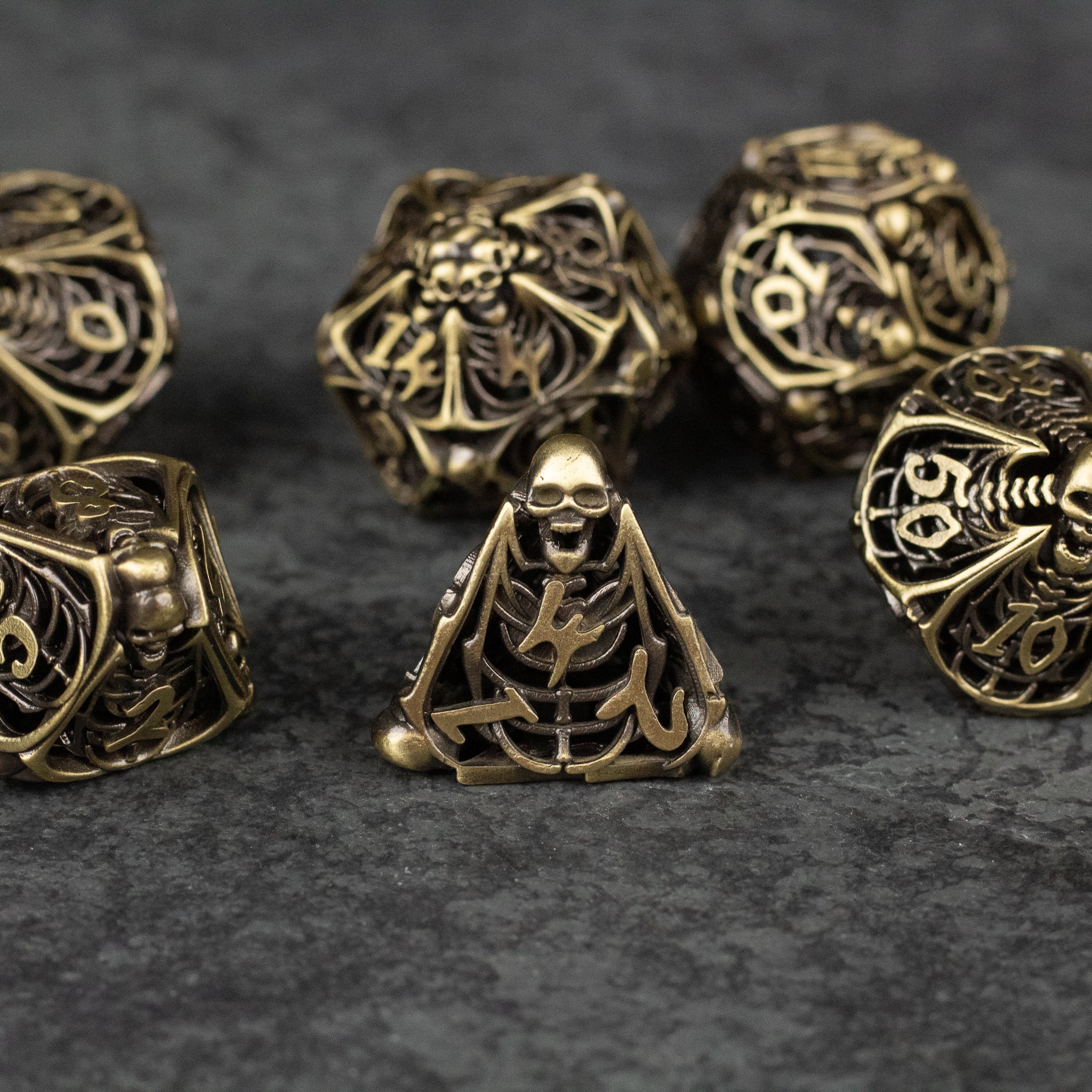 Bone Crypt Dice Set - Bronze-colored metal dice set with hollowed designs featuring skulls and bone motifs on a dark textured surface.