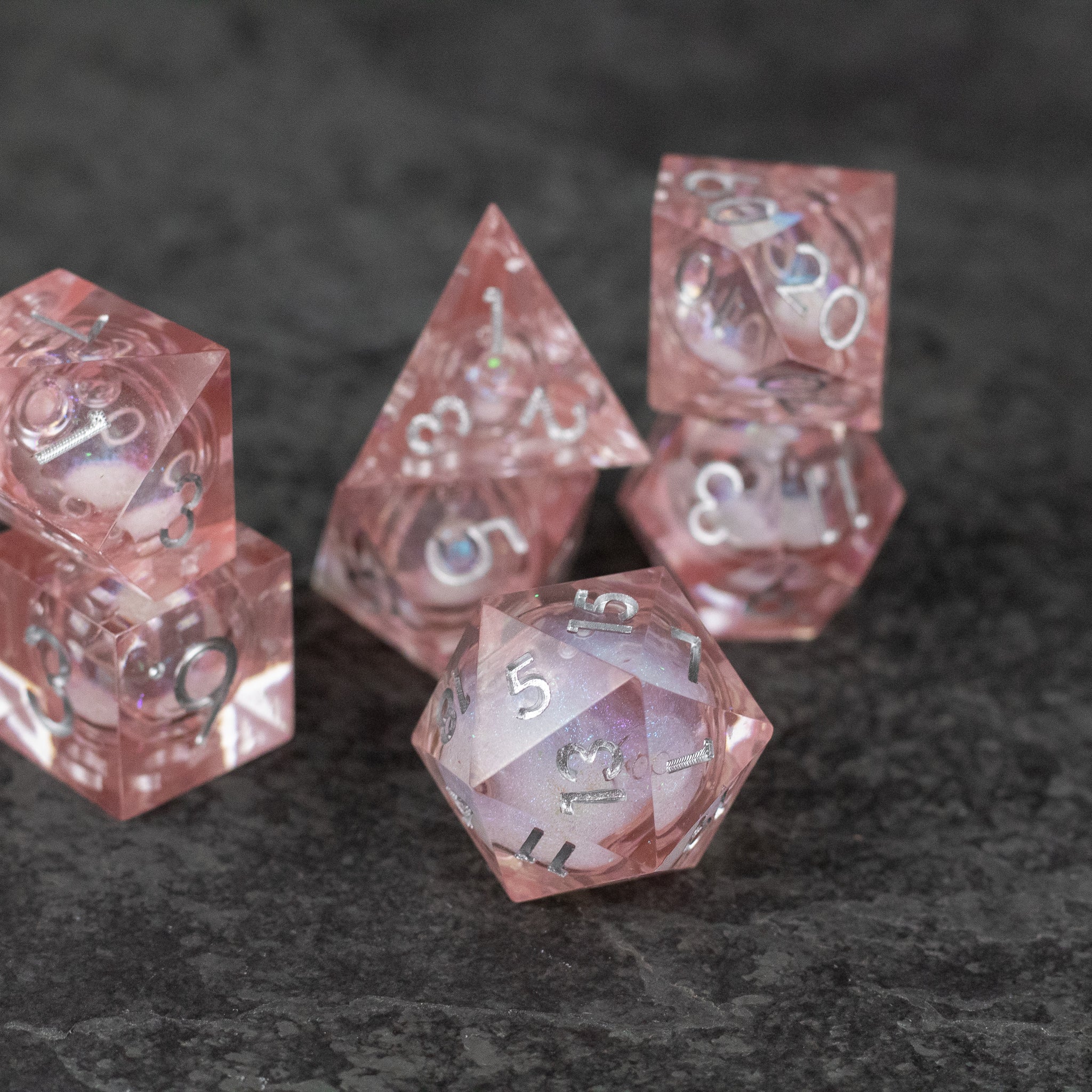 Fairy Dust - Liquid Core Dice Set - Set of six pink translucent sharp-edged dice with liquid core and white numbers on dark stone surface.
