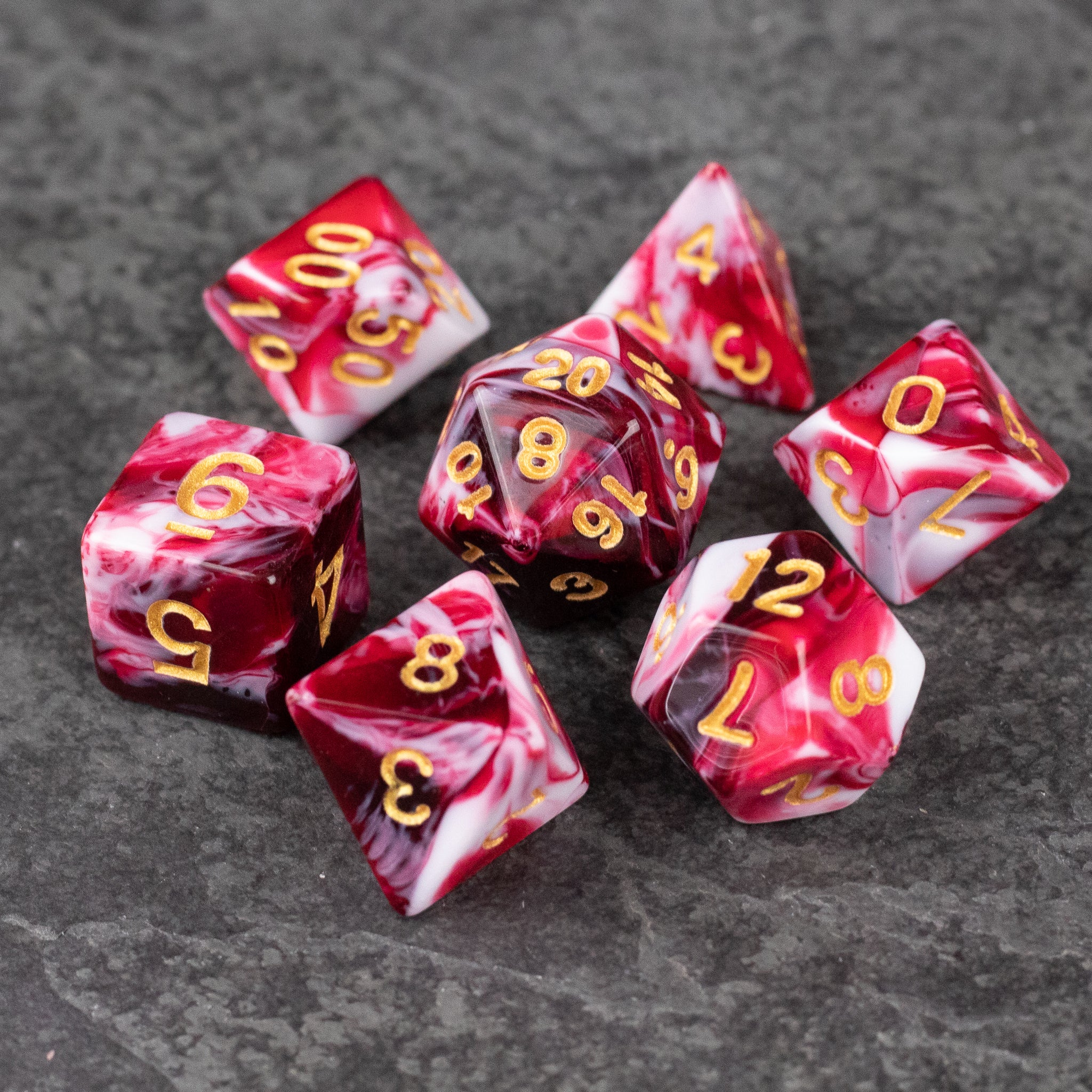 Red and White Marbled Dice Set - Seven red and white marbled acrylic dice with gold numbers on a dark textured surface.