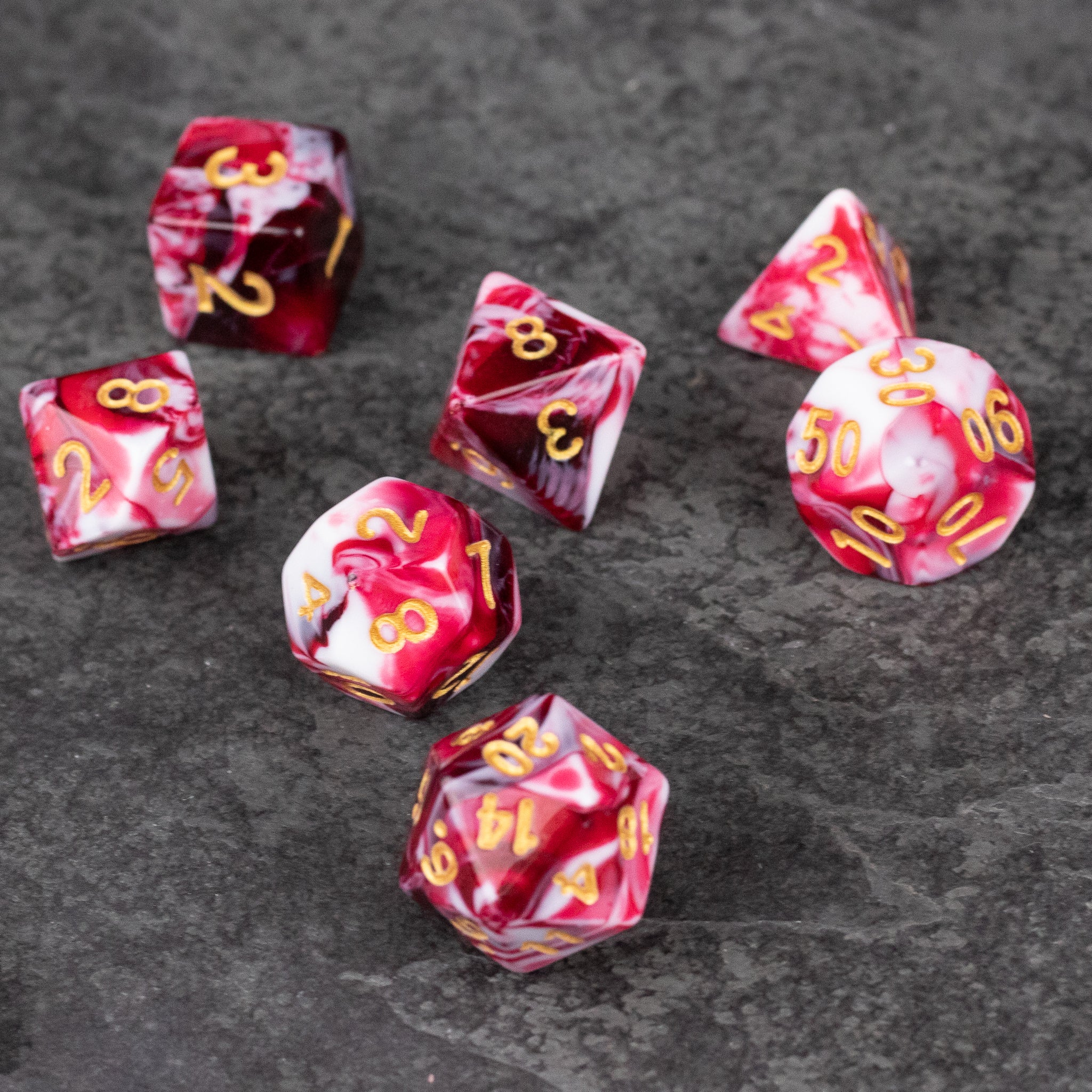 Red and White Marbled Dice Set - Seven red and white marbled acrylic polyhedral dice with gold numbers on a gray surface.